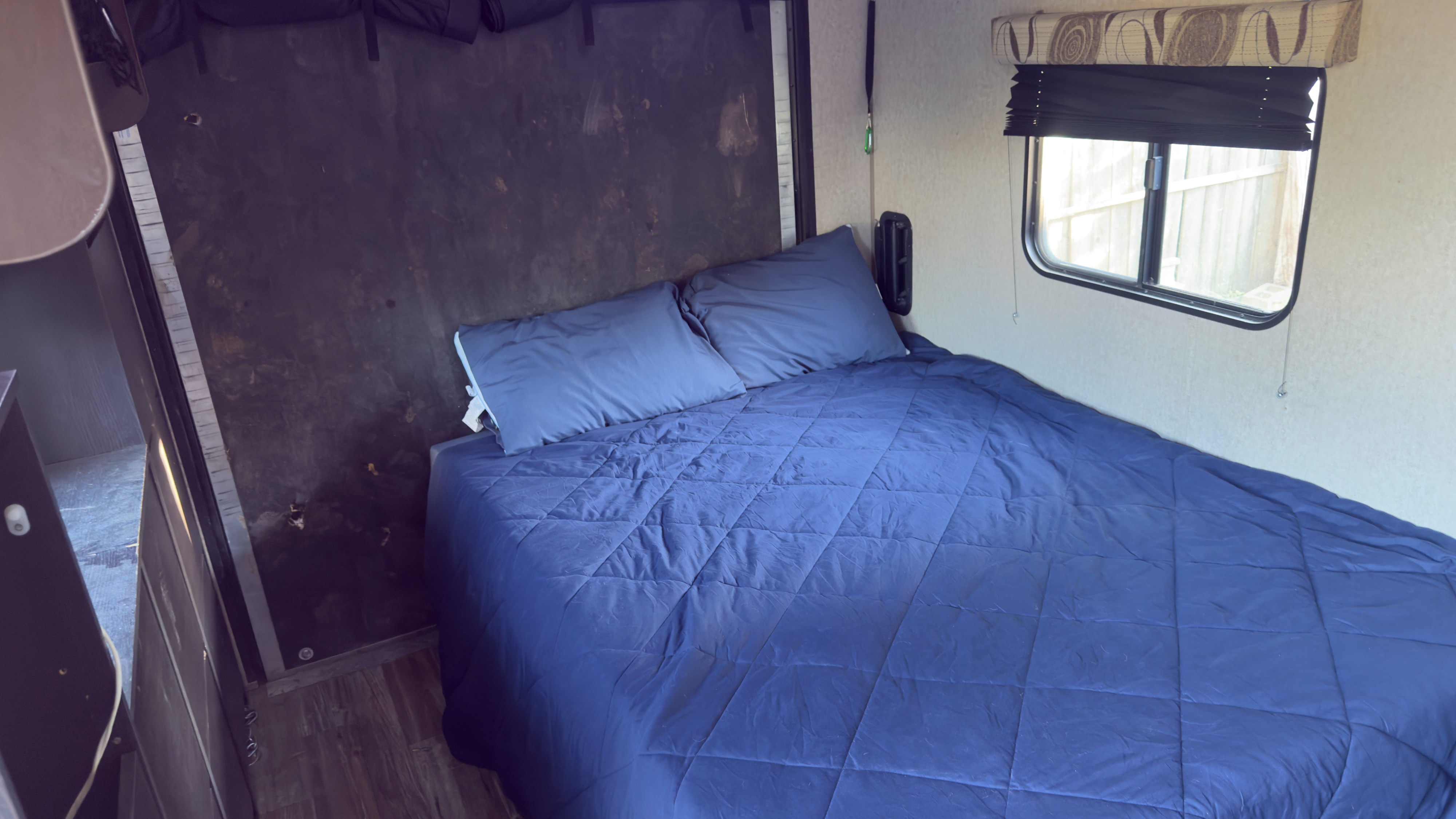 This RV rental's bedroom shows a blue bed, pillows, dark wall, and a window with blinds, offering a cozy sleeping space.. Forest River Cherokee Grey Wolf Toy Hauler 2016