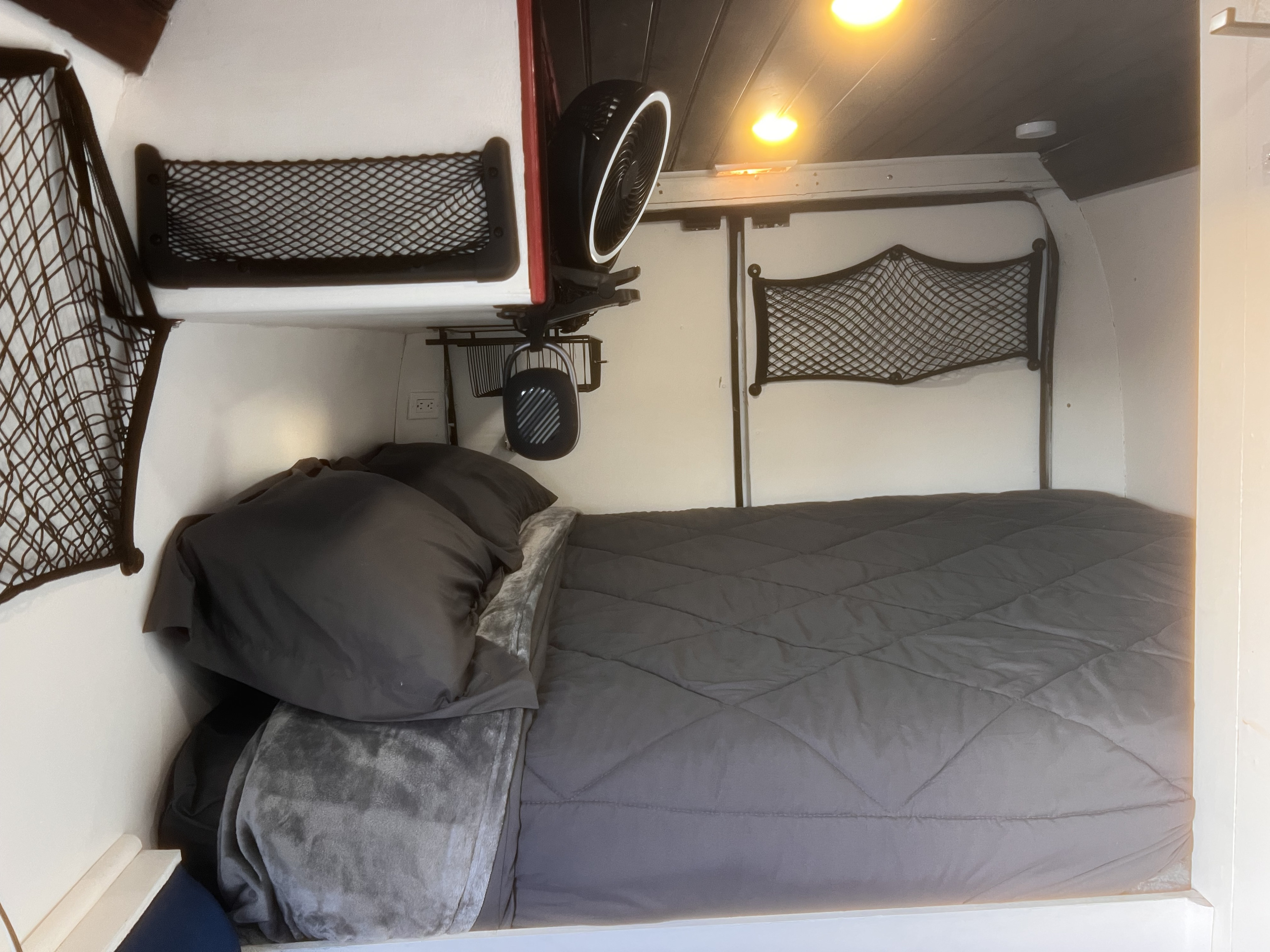Cozy RV rental bed area with gray comforter, pillows, fan, lights, and mesh storage nets for camper travel.. Ram Promaster 2500 2019