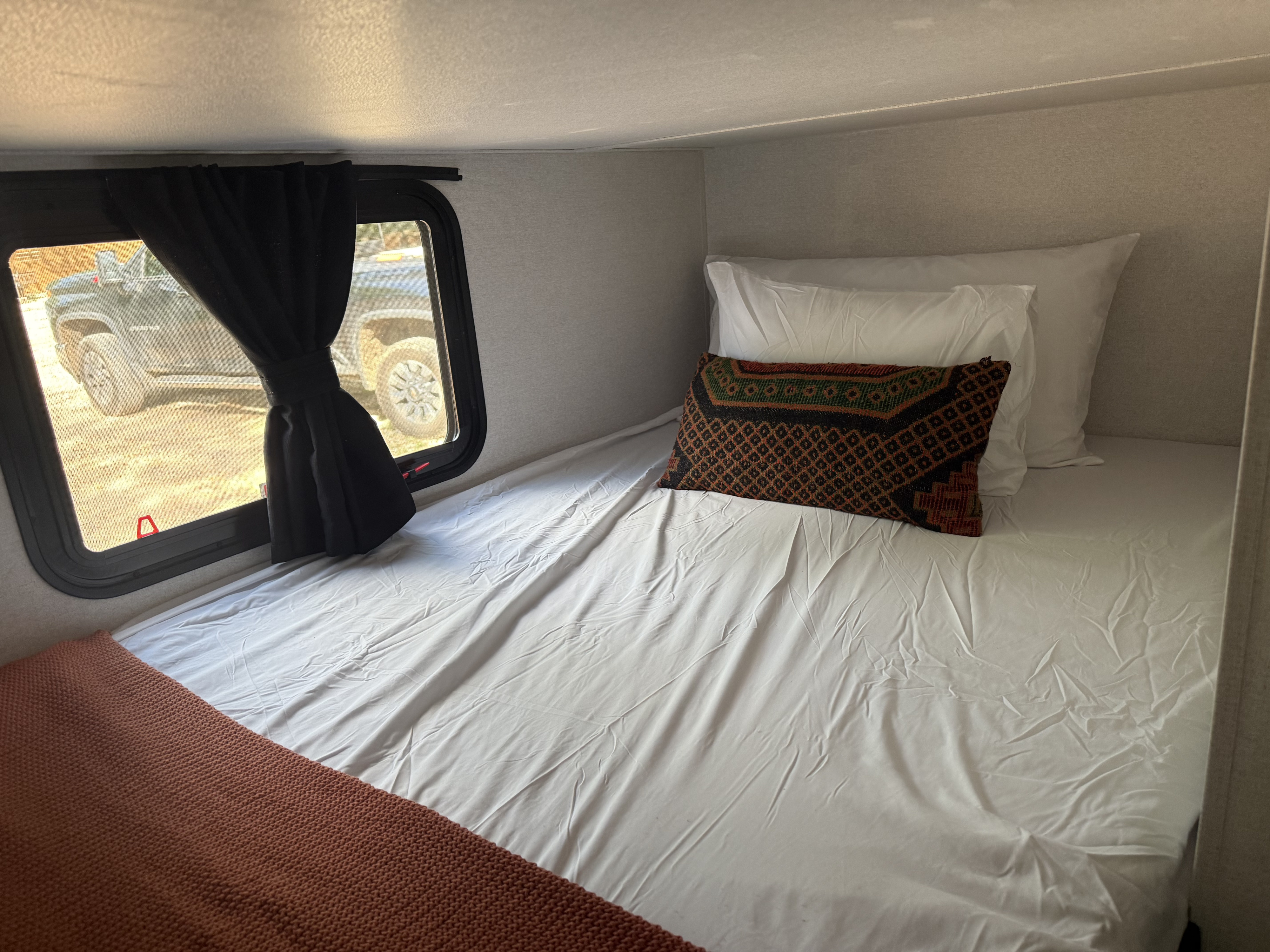 Inside an RV rental: a neat bed with white linens, decorative pillow, and a window offering a view of a parked truck.. Jayco Jay Flight 260BH 2026