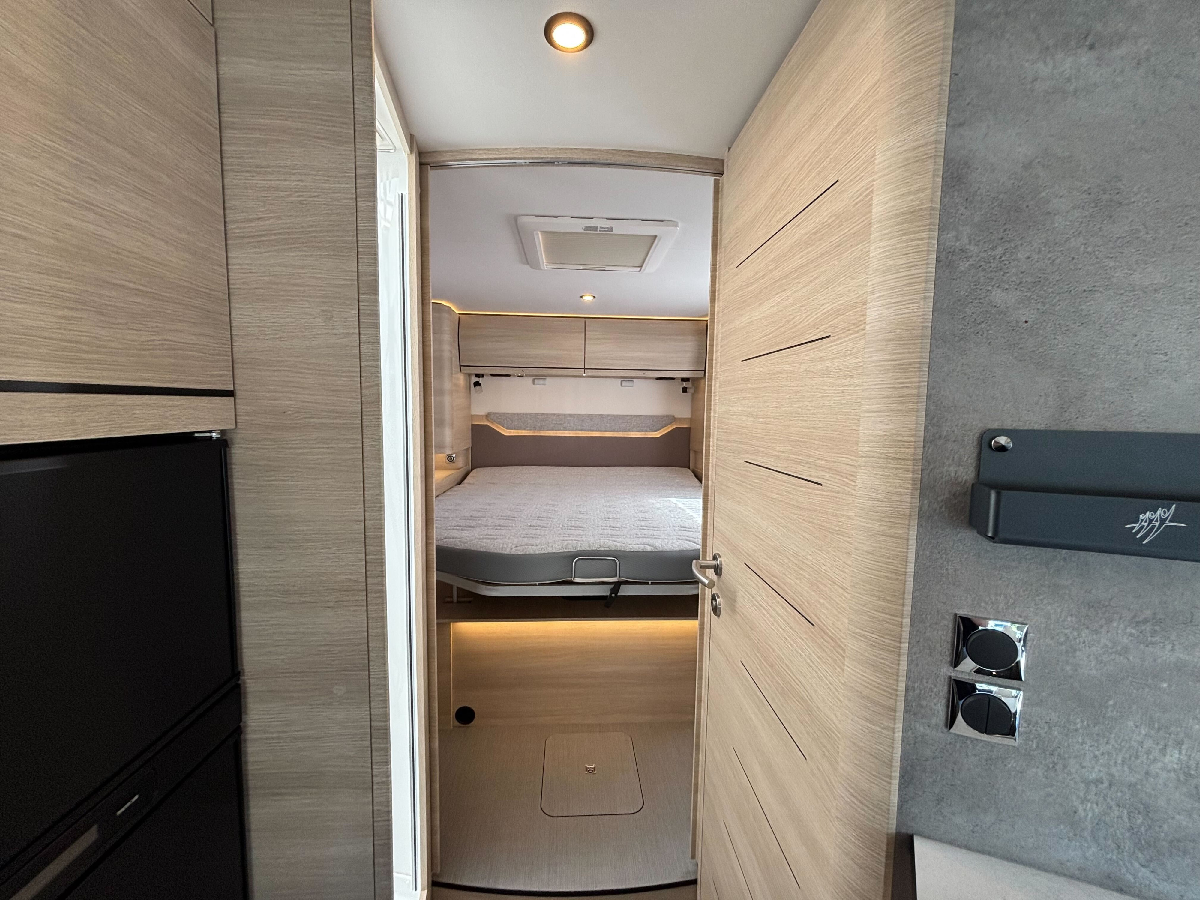 Step inside this modern RV rental! View into a bright bedroom from the living area, featuring a cozy bed, overhead storage, and light wood interior.. RAPIDO 896 F 2025