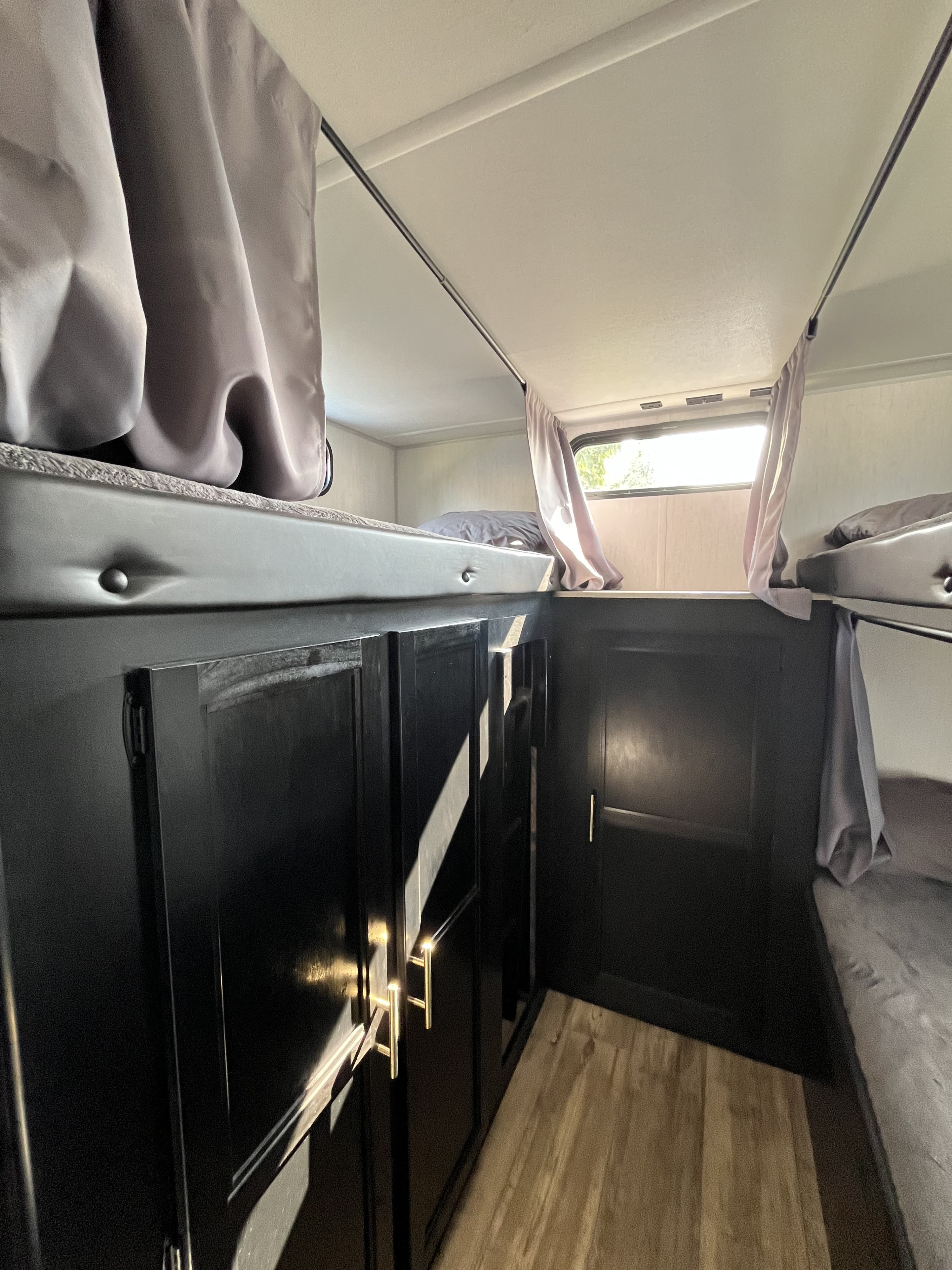 RV rental bunk area: black cabinets with gold pulls, comfy gray bedding, and curtains for privacy, illuminated by a window.. Forest River Cherokee Grey Wolf 2022