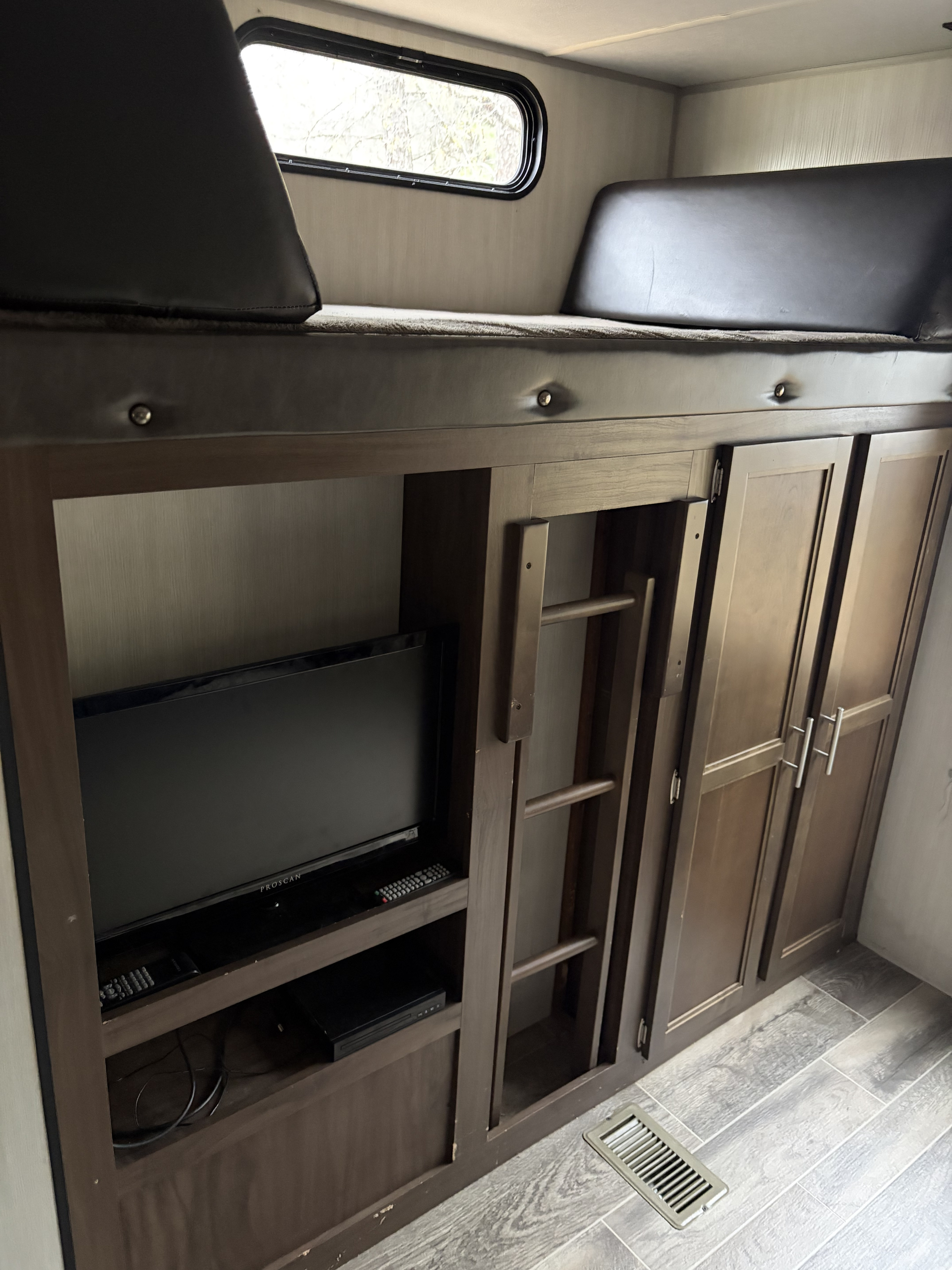 RV rental bunk area: wood-toned storage unit with TV, ladder, cabinets, and a black-cushioned upper bunk with window.. Forest River Cherokee 2019