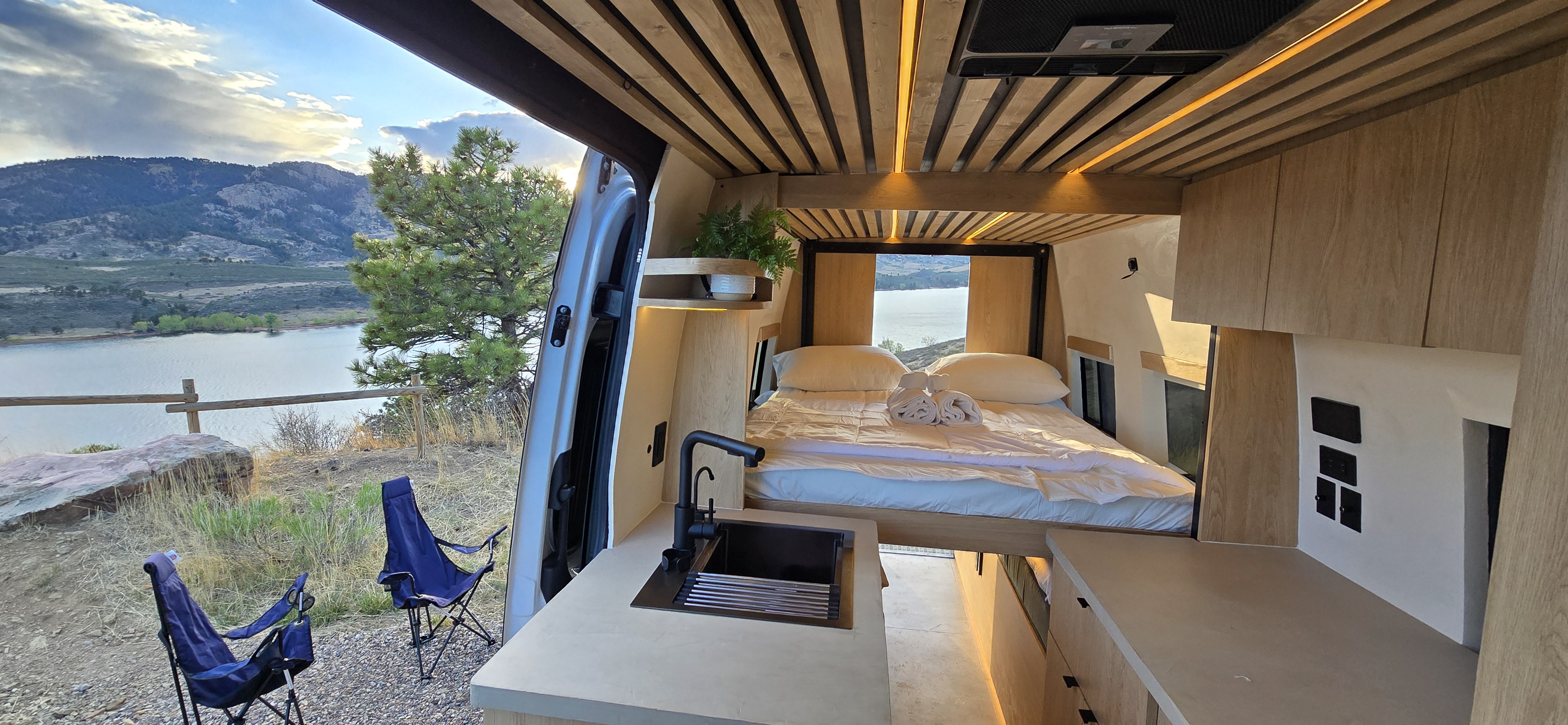 RV rental camper van interior featuring a comfy bed, kitchen sink, and a scenic lake and mountain view from the open door.. Mercedes Mercedes Sprinter Adventure Van 2025