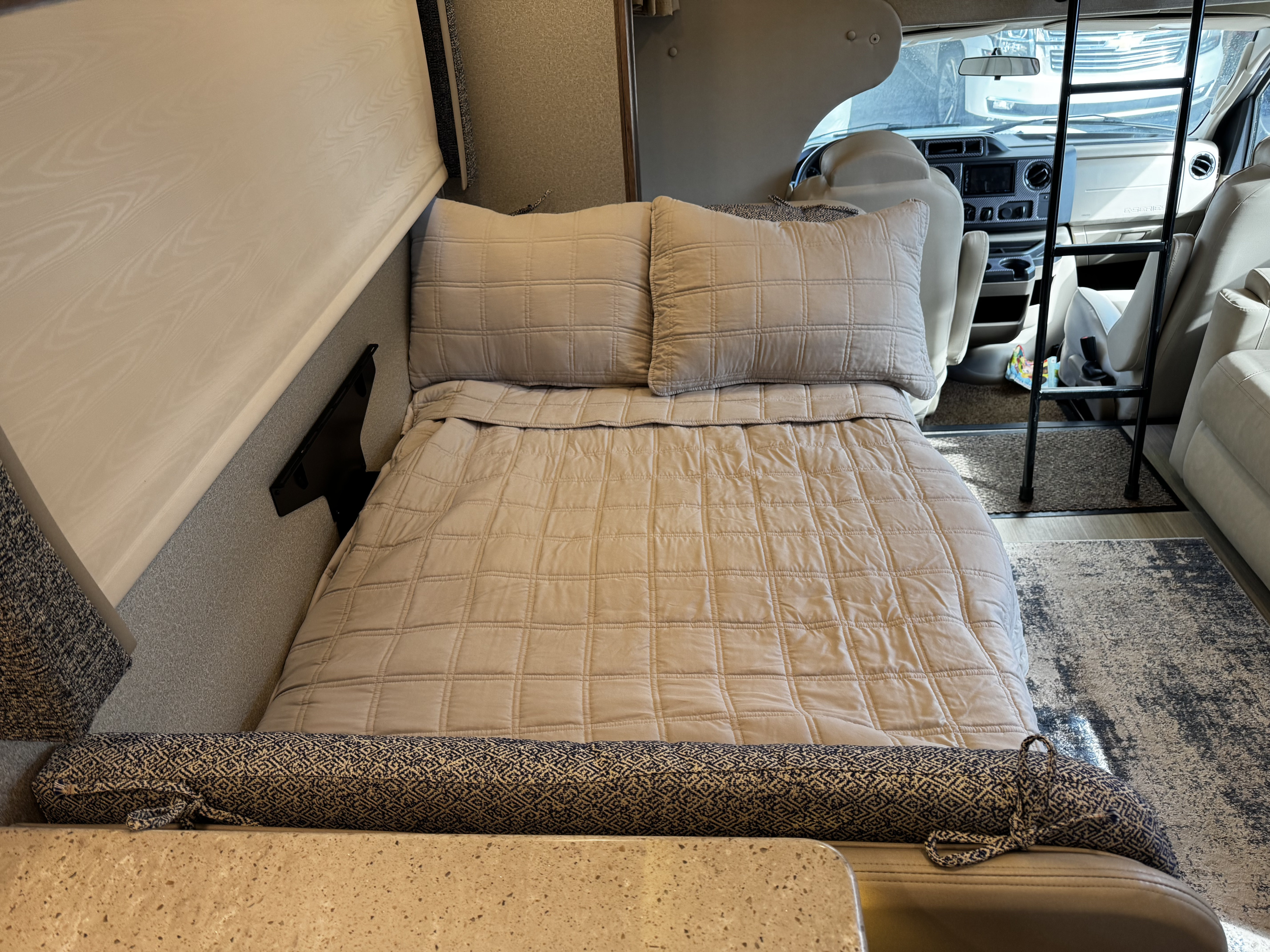 Inside a comfortable RV, showing a neatly made bunk bed with neutral bedding and a view of the driver's cab. Ideal for an RV rental.. Jayco Greyhawk Prestige 2020
