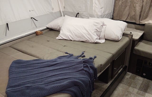Relax in this RV rental's comfy sleeping area. Gray cushions, white pillows & a blue blanket create a cozy spot in this pop-up camper.. Forest River Rockwood Freedom 2021