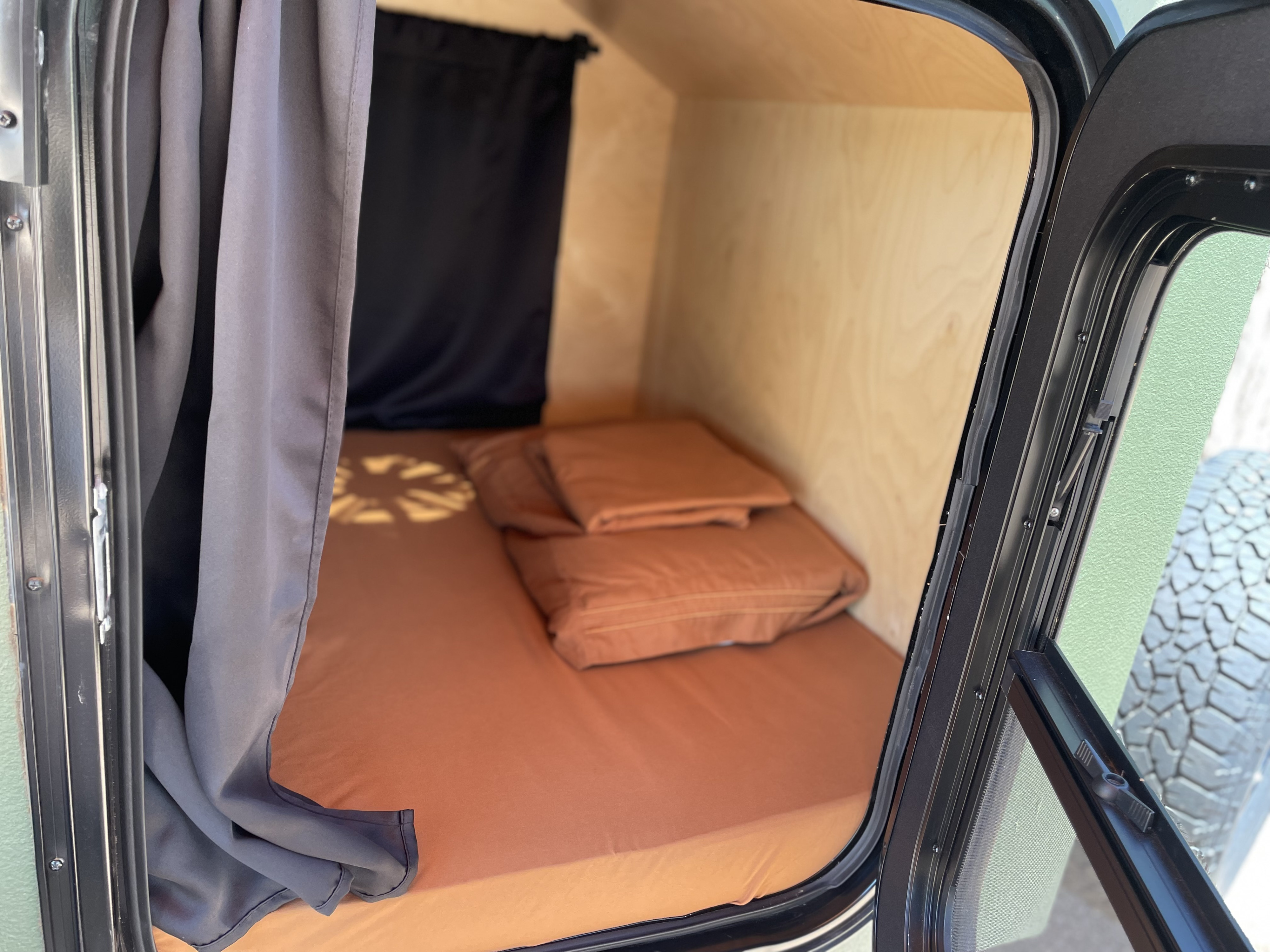 Cozy rv rental sleeping area with orange bedding, wooden walls, and privacy curtains. Ideal for compact travel.. Off Grid Trailers Off Grid Trailers Trailer 2022