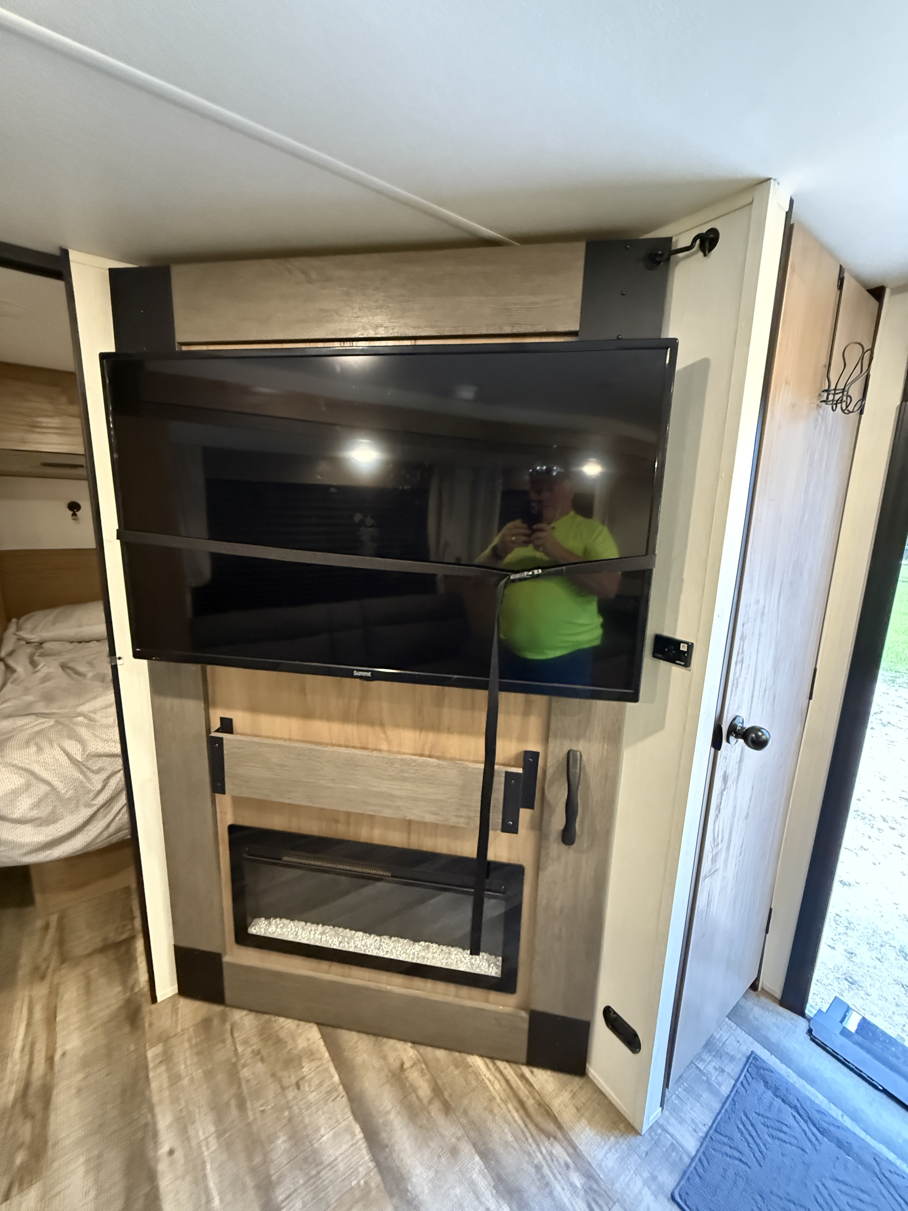 Interior of an **rv rental** camper: TV over a fireplace, partial bedroom, and a person's reflection in the screen.. Forest River Campsite Reserve 2025