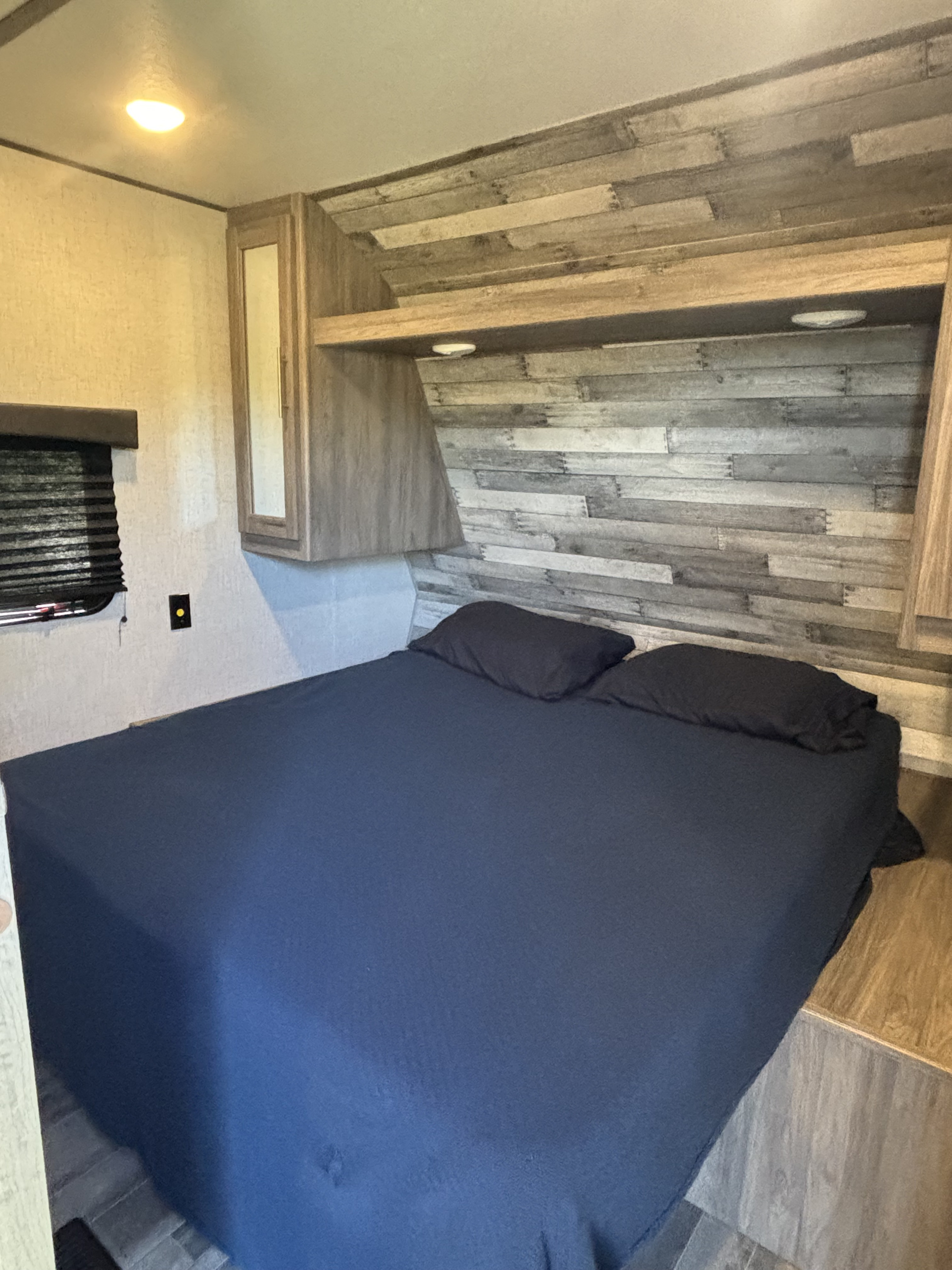 View inside an RV rental bedroom: bed with blue bedding, wood-planked headboard, overhead storage, and window.. Keystone RV Zinger 2022