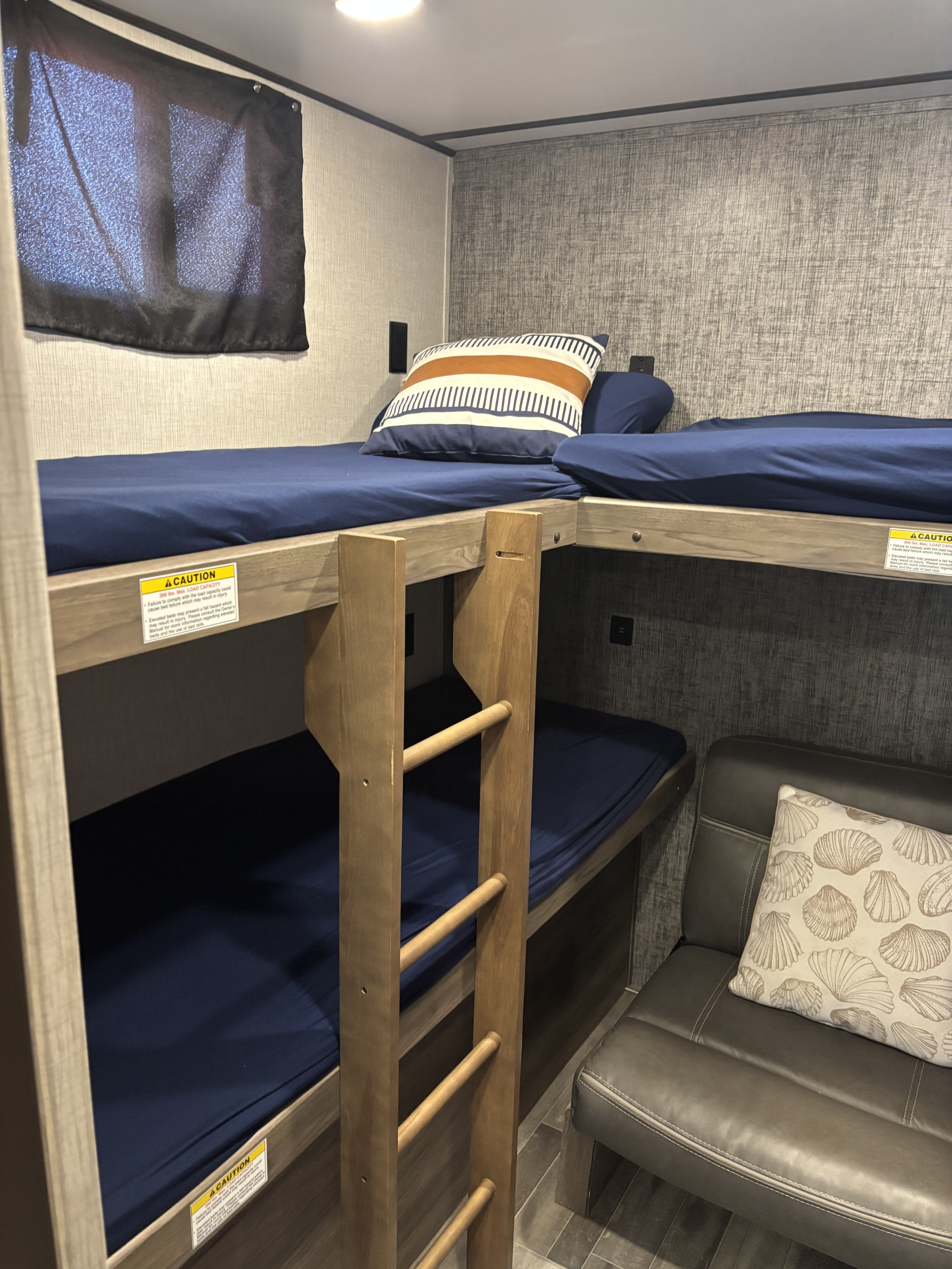 RV rental bunk beds feature navy sheets, a wooden ladder, and a striped pillow. Below, a plush seat with a seashell pillow and window.. Keystone RV Zinger 2022