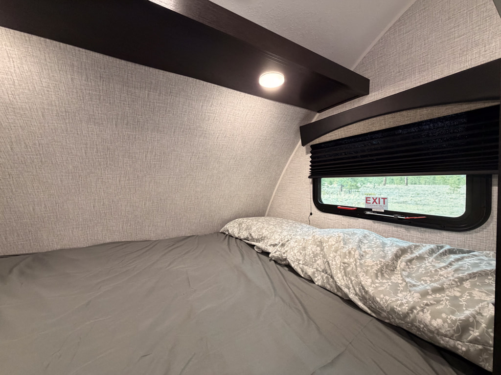 Cozy RV rental bedroom with gray bedding, a patterned pillow, and a window showing an "EXIT" sign to greenery.. Braxton Creek Bushwhacker Plus 2022