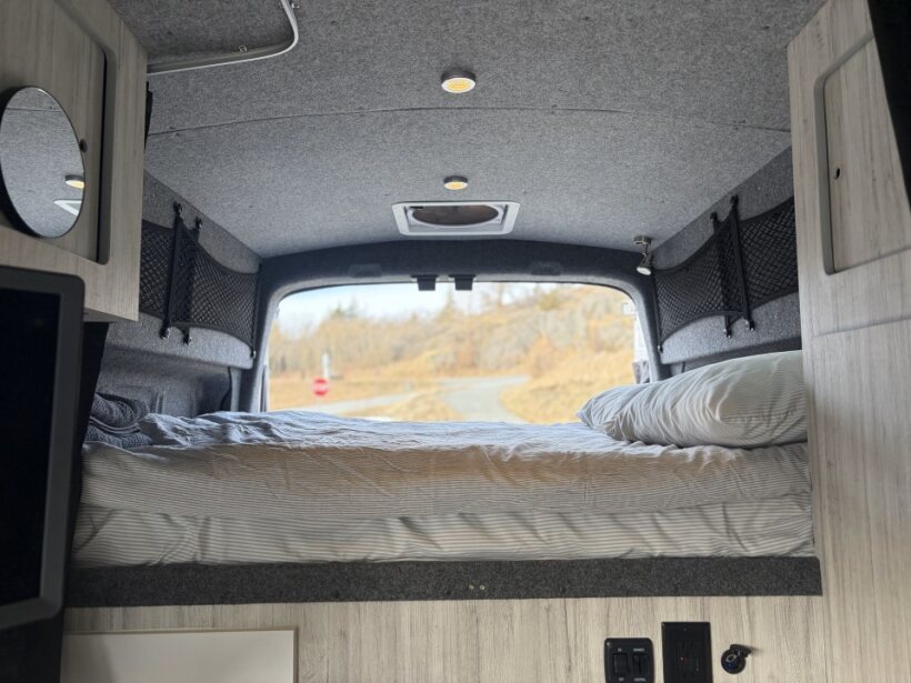 This RV rental offers a cozy camper van bed facing the rear window, overlooking a scenic road and trees for mobile living.. Ford Transit 2022