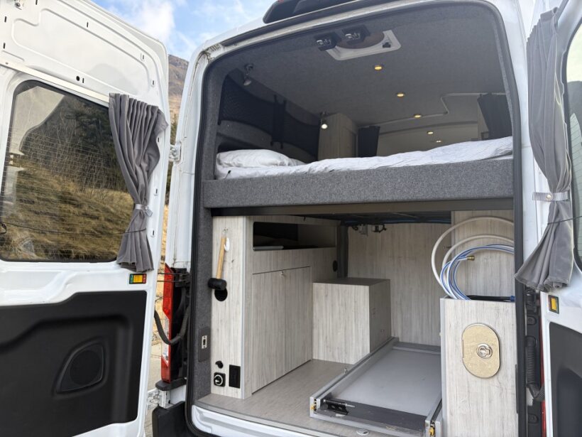This inviting RV rental interior features an elevated bed, pull-out storage, and outdoor shower hookup, ready for adventure.. Ford Transit 2022