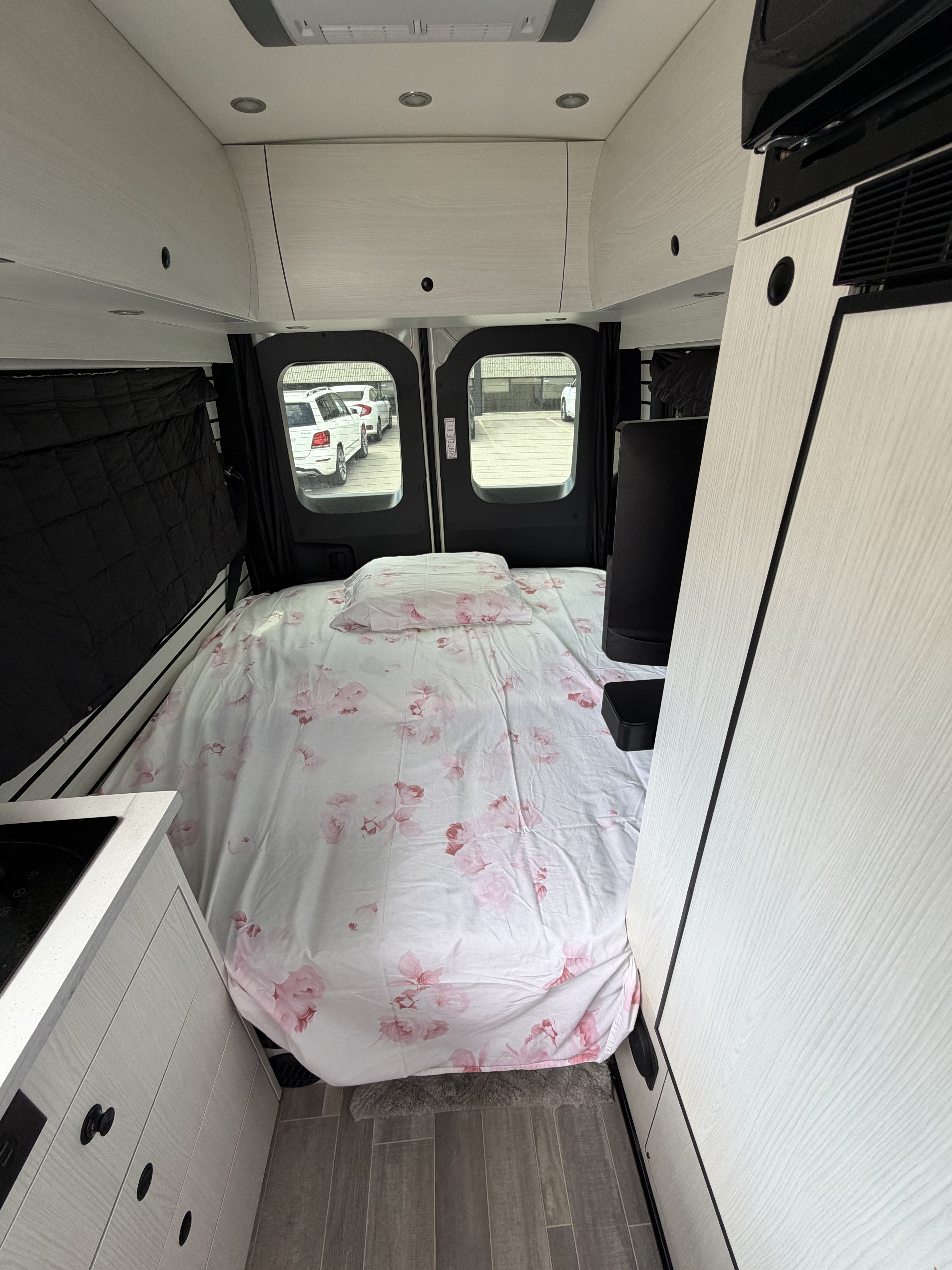 RV rental interior: cozy campervan bed with floral sheets, rear windows, compact kitchenette, and light overhead storage.. Pleasureway Ascent 2026