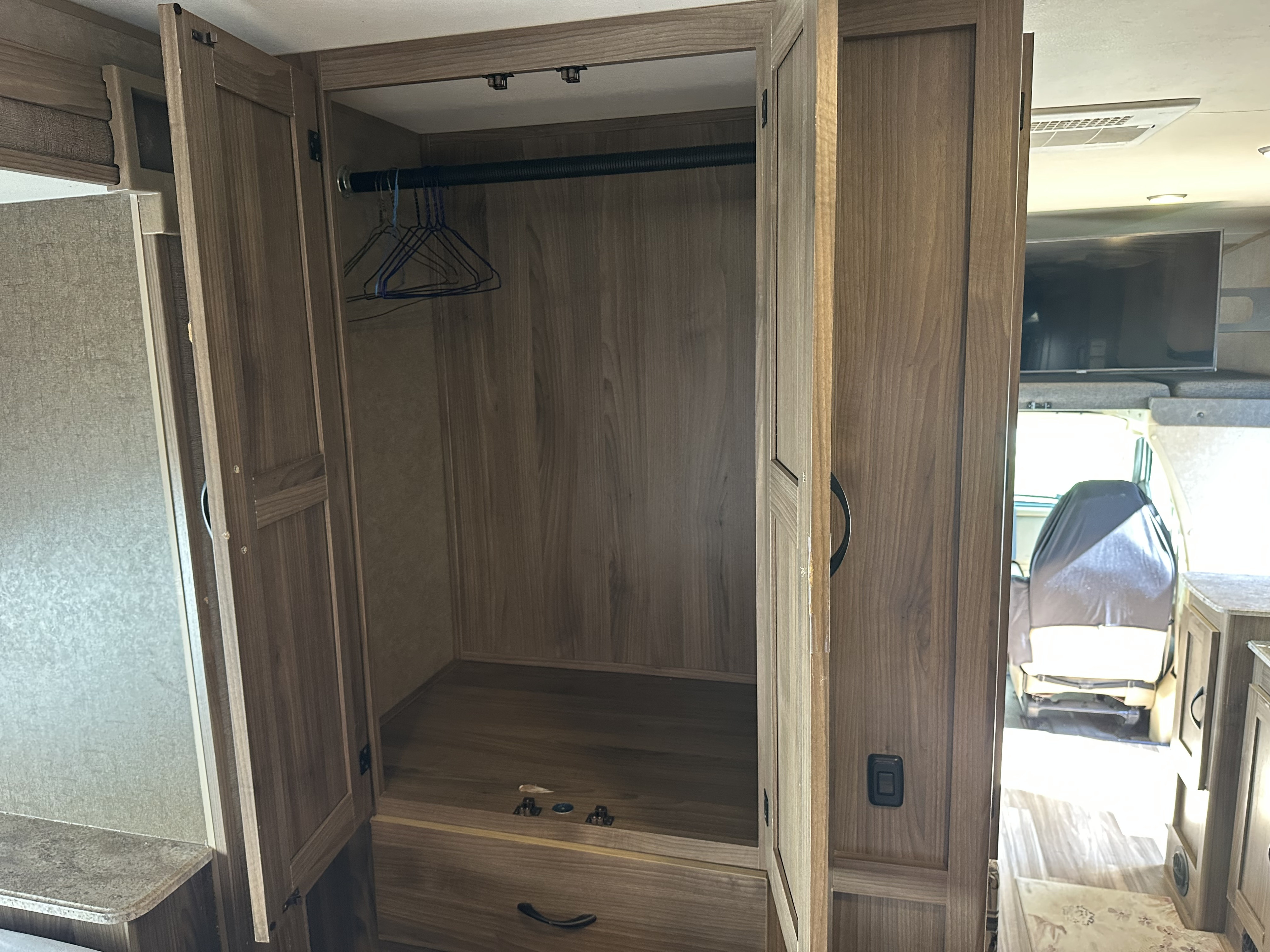 RV rental wardrobe: open wood closet with empty hangers & drawer. View into RV cockpit with a covered seat and TV.. Coachmen Freelander 2015