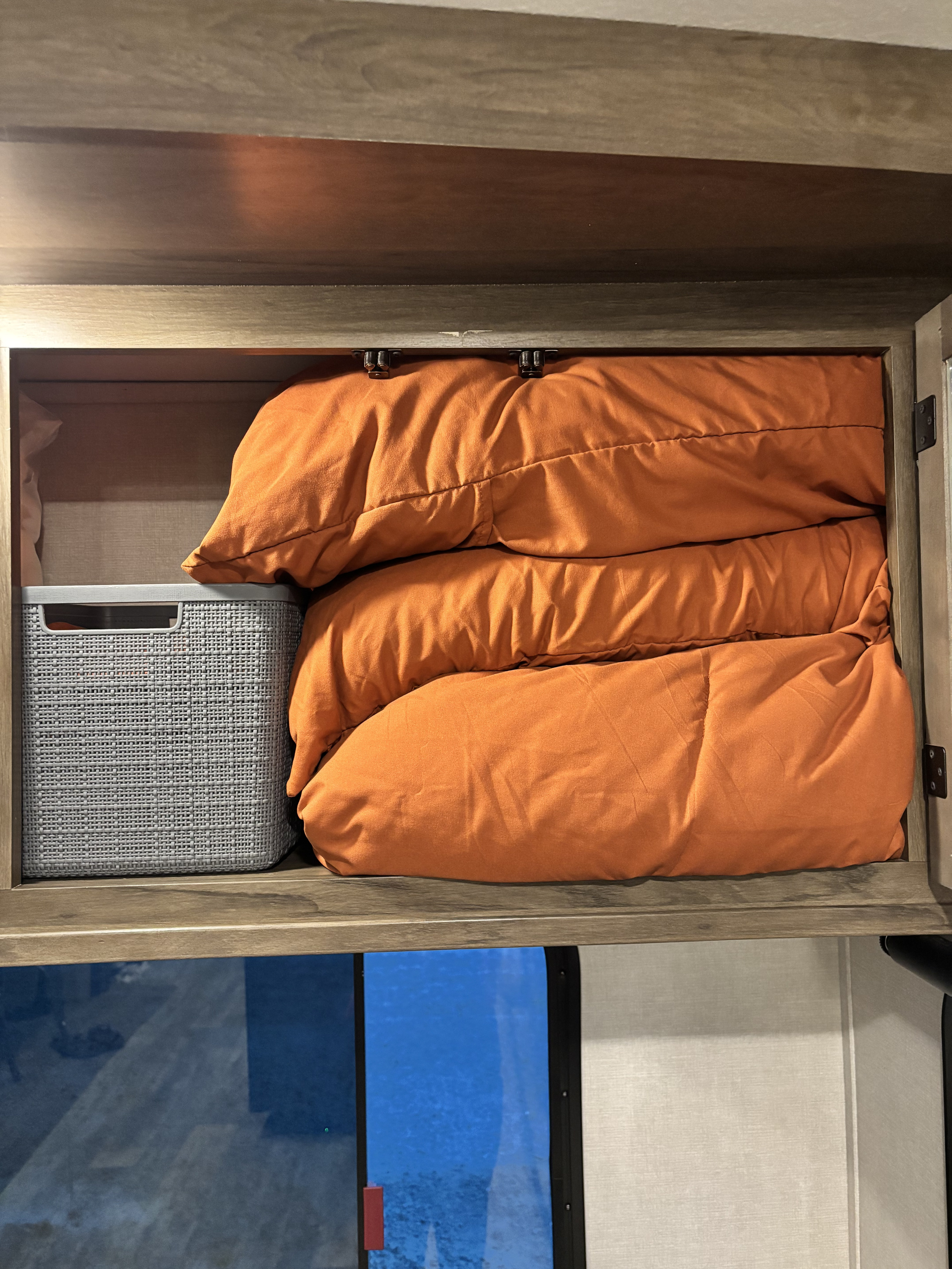 RV rental overhead cabinet with neatly folded orange comforters and a gray woven basket, showcasing organized storage.. Forest River Sanibel 2021