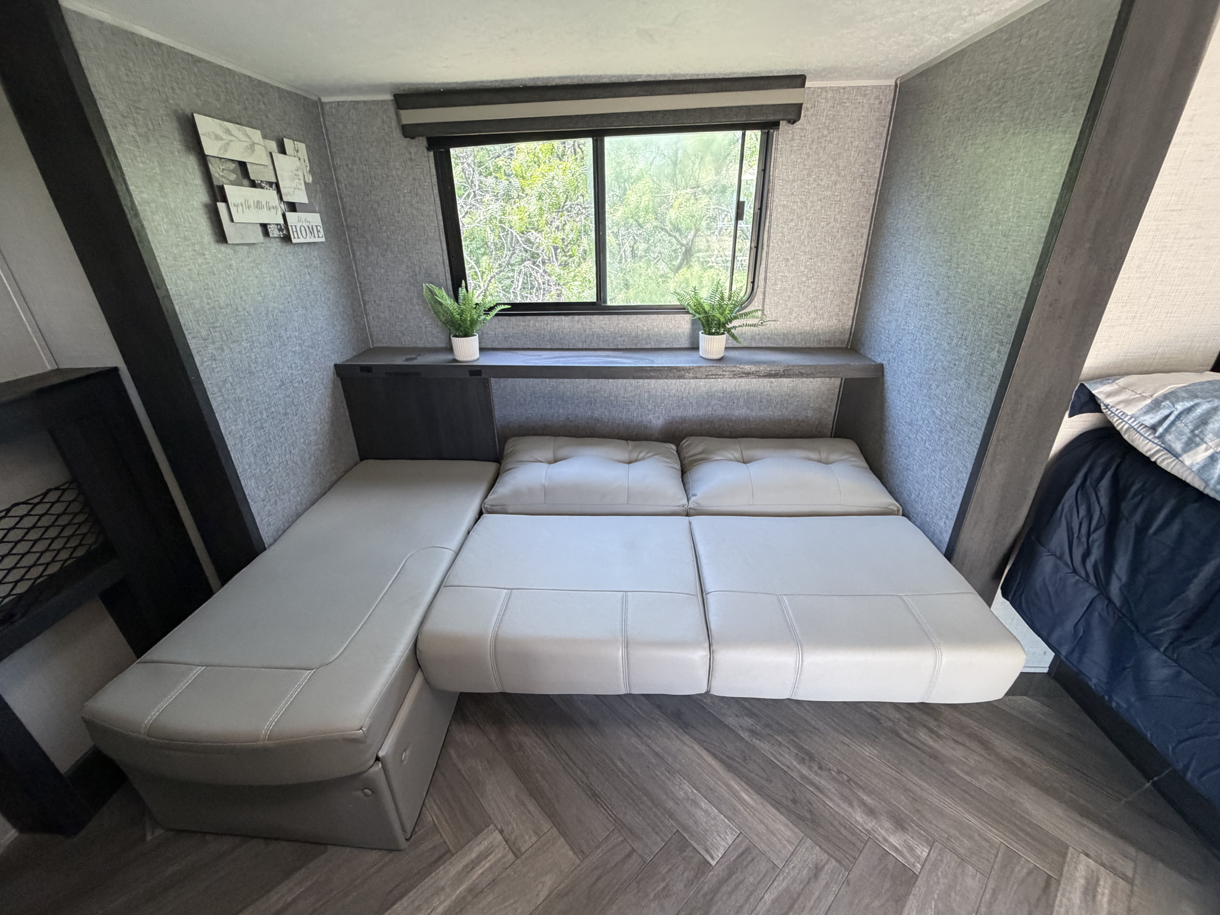 Compact RV rental interior: versatile sofa/bed with gray upholstery, window view, and small plants on a shelf. A functional living/sleeping space.. Forest River RV Ozark 2021