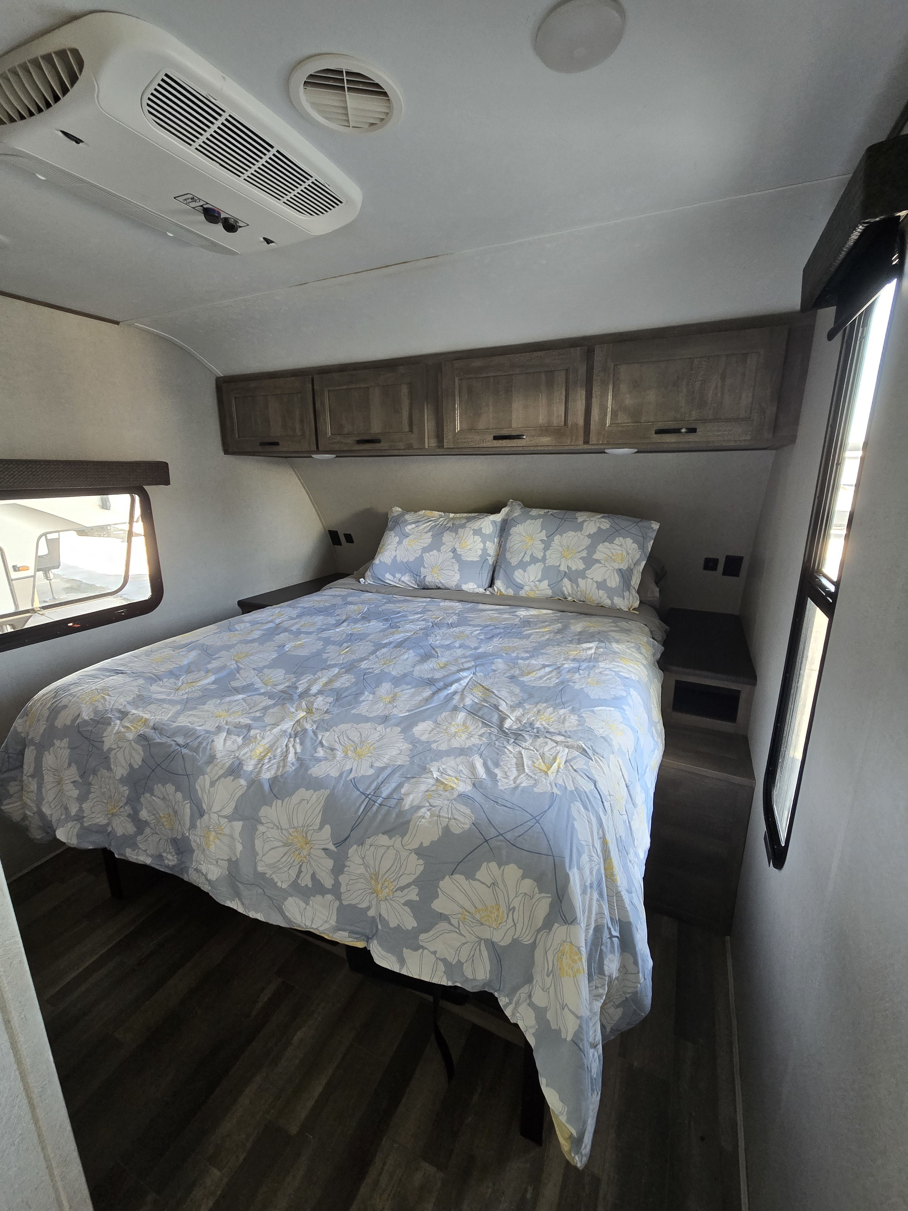 Comfortable RV rental bedroom featuring a queen bed with blue floral bedding, overhead cabinets, windows, and AC unit.. Forest River Vibe 2020