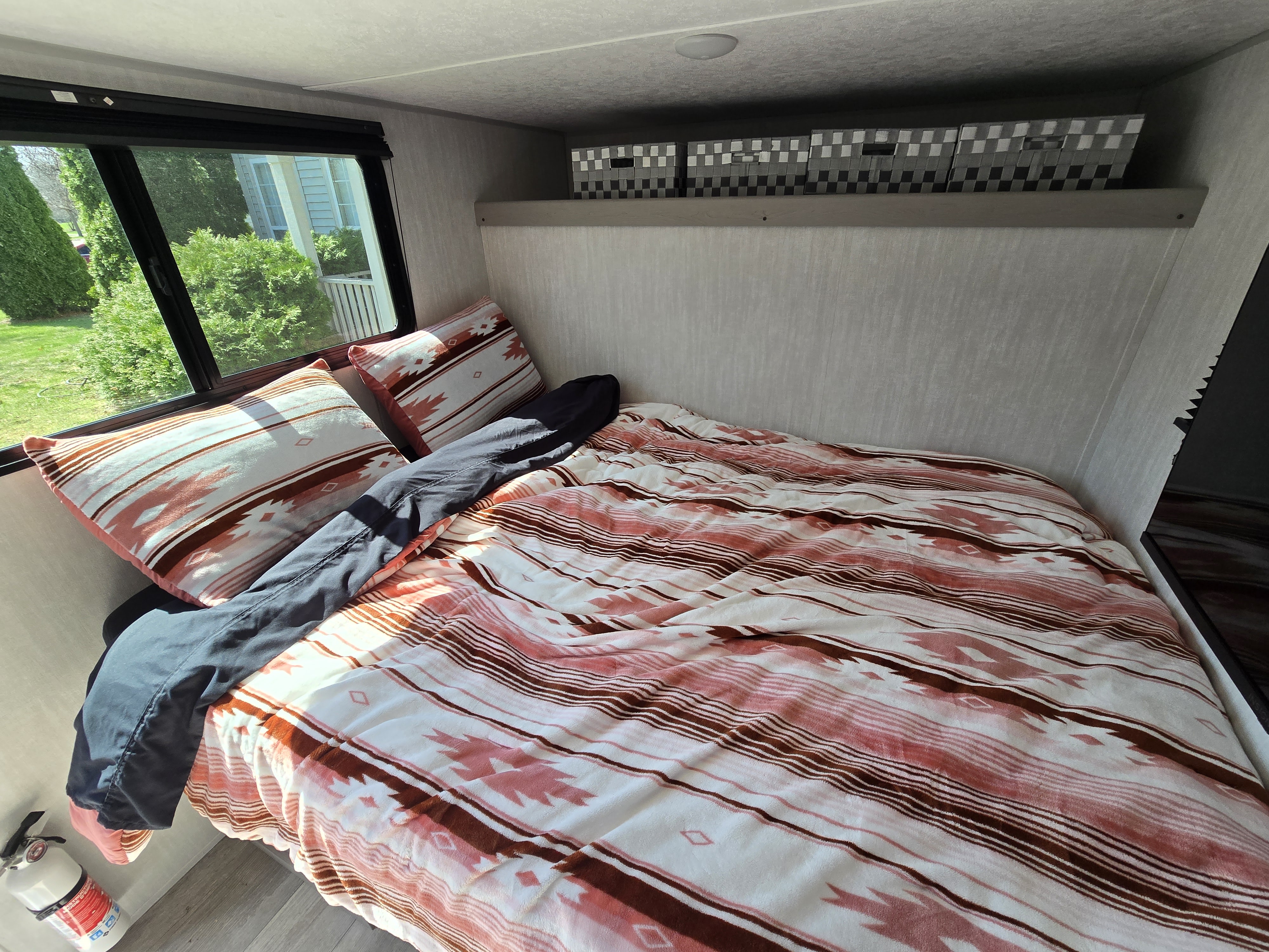 RV rental bedroom showing patterned bedding, a window with outdoor views, overhead storage bins, and a fire extinguisher.. Clipper 3K 2025