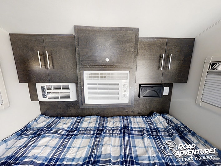 Cozy RV rental bedroom with a plaid bed, overhead cabinets, and dual AC units, perfect for comfortable road adventures.. nuCamp T@G XL 2026