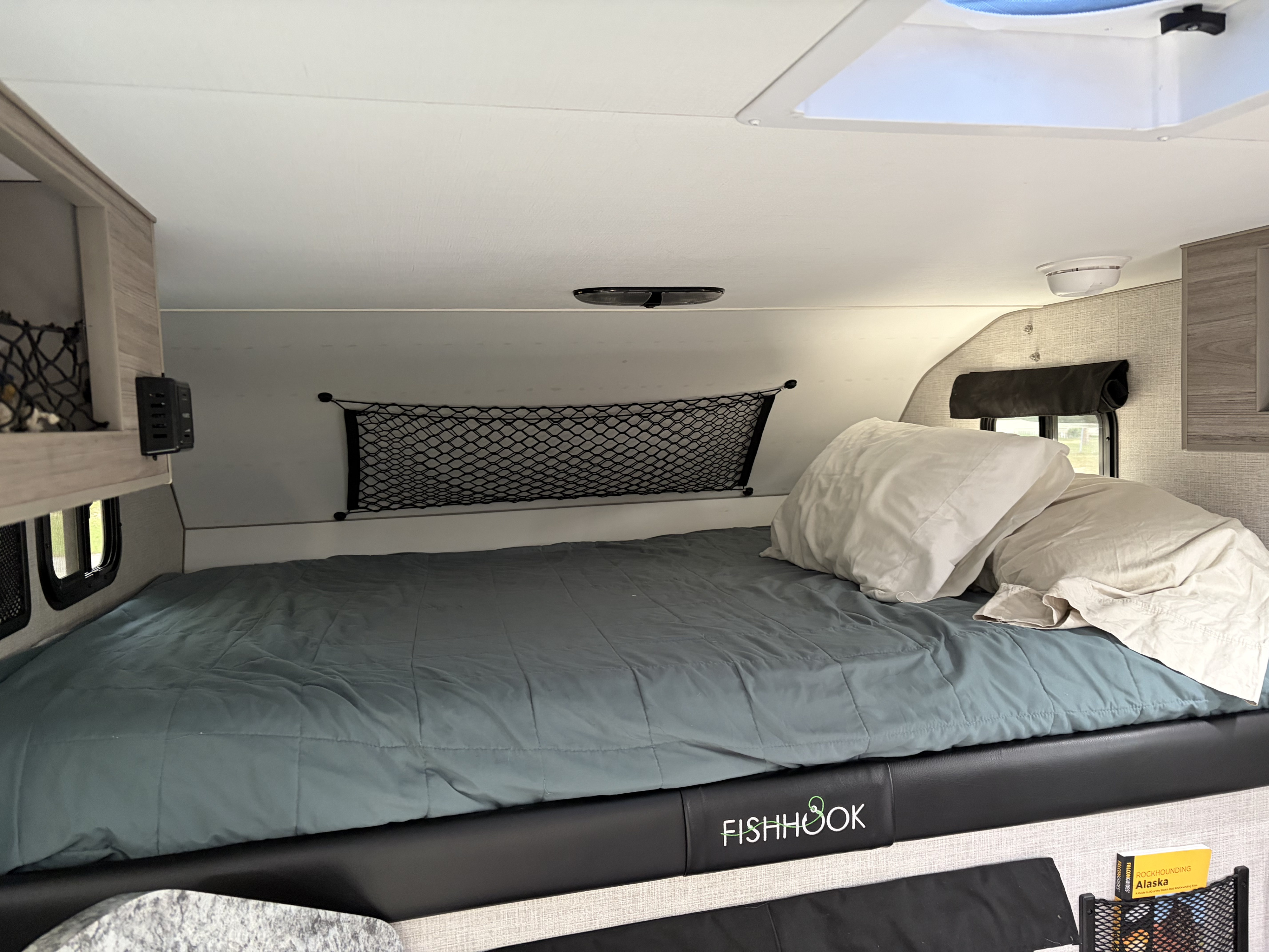 RV rental interior featuring a comfy bed with "FISHHOOK" detail on the frame, storage nets, and a skylight.. Custom Camper 2025