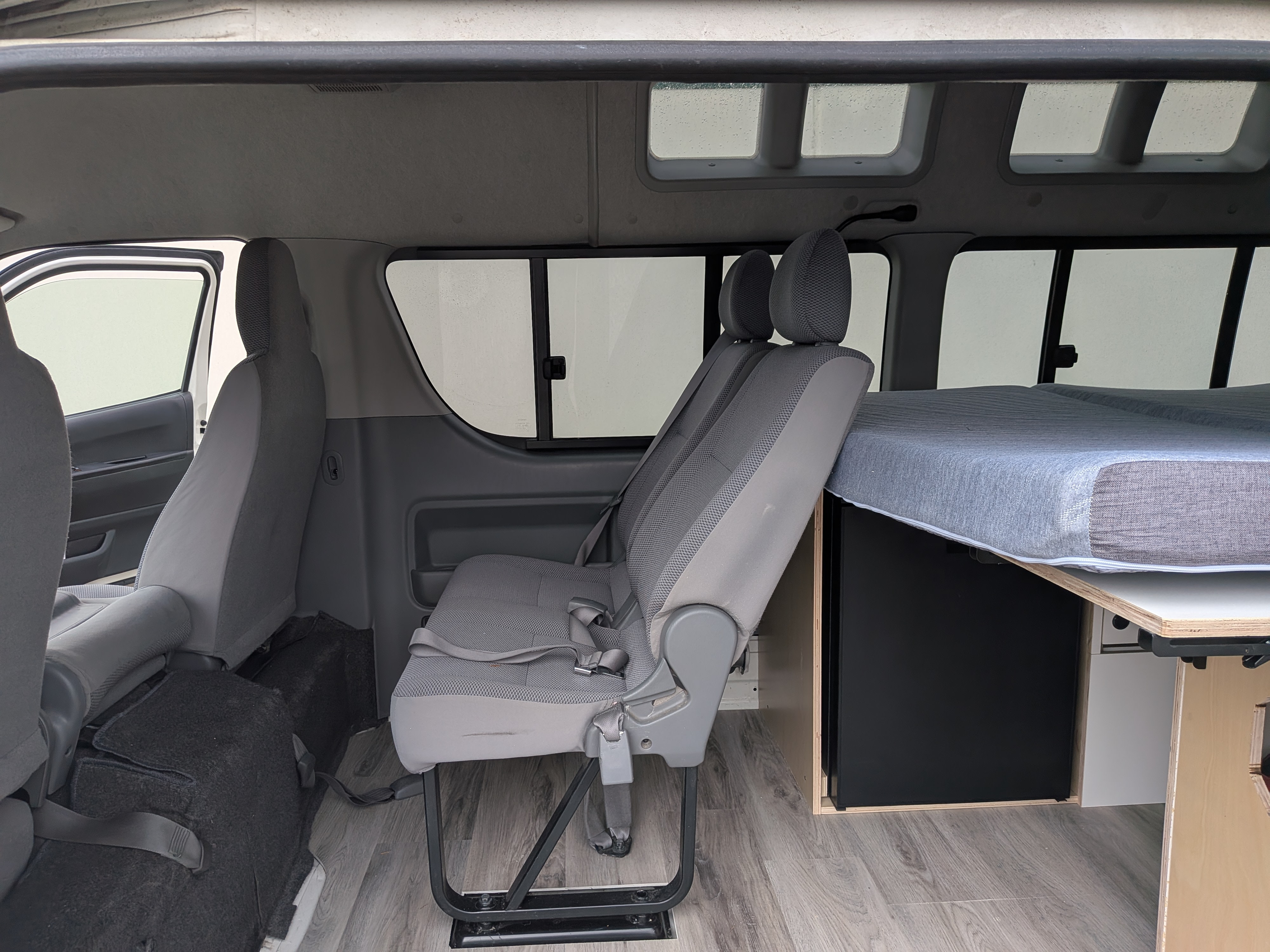RV rental camper van interior: grey front & middle seats, wood-look floor, and a bed setup with storage cabinetry.. Toyota Hiace 4WD 2009