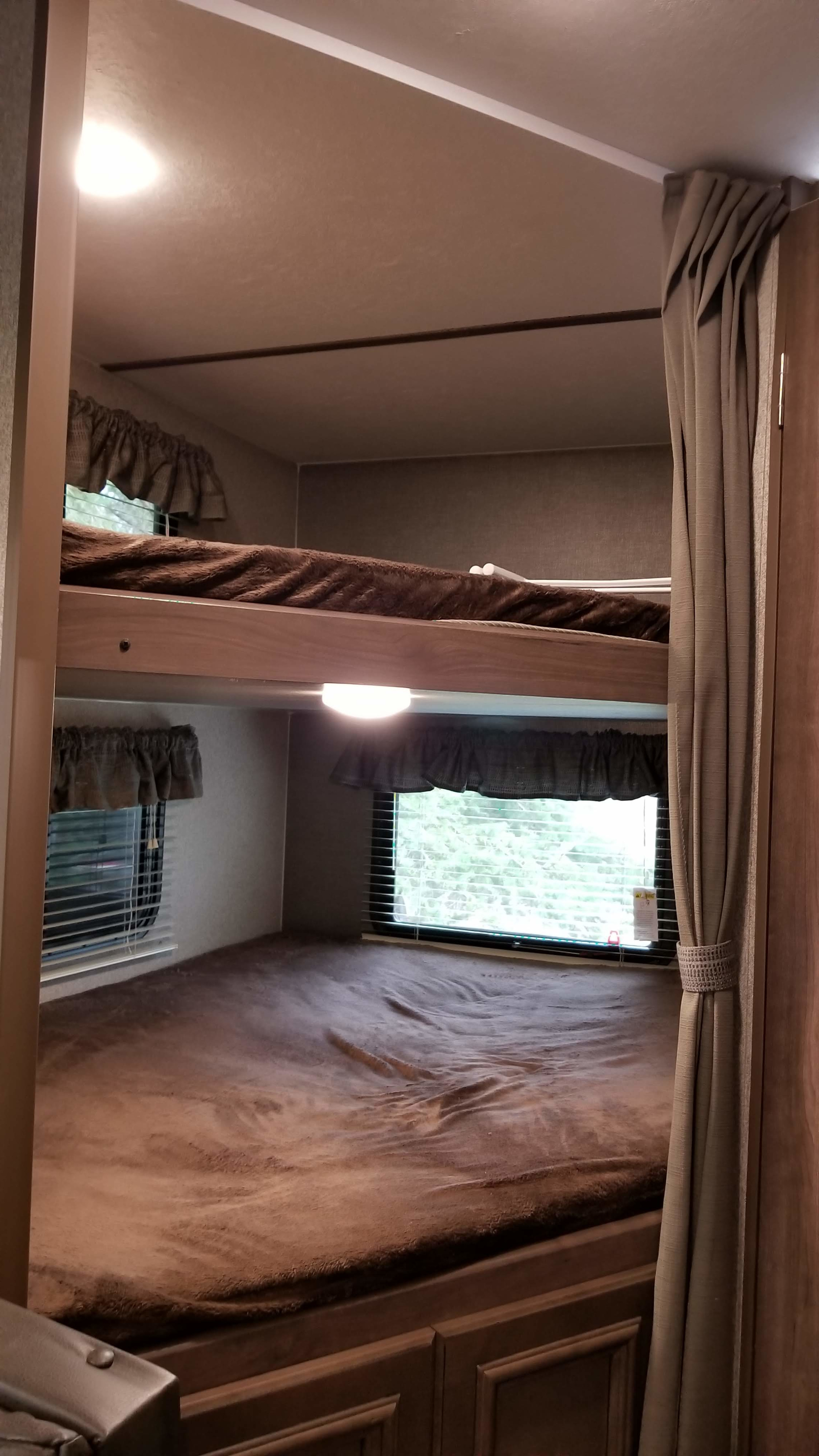 This RV rental bunk bed area features cozy brown blankets, windows with blinds, and a curtain, ideal for a comfortable stay.. Keystone RV Passport Ultra Lite 2019