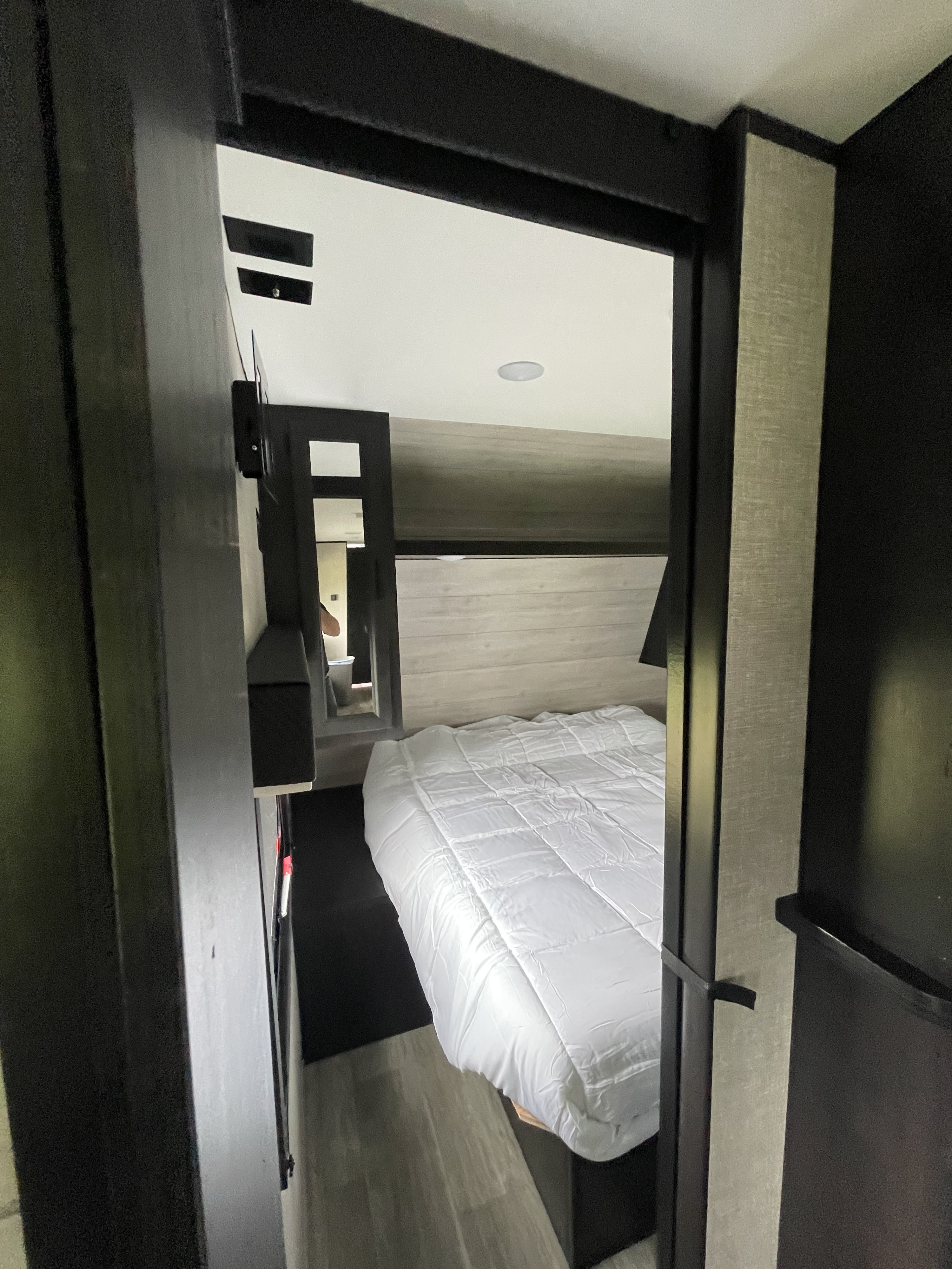 RV rental bedroom with a clean white bed, dark gray walls, and light paneling. A view into an adjacent area is seen through a doorway.. Jayco Jay Flight 2022
