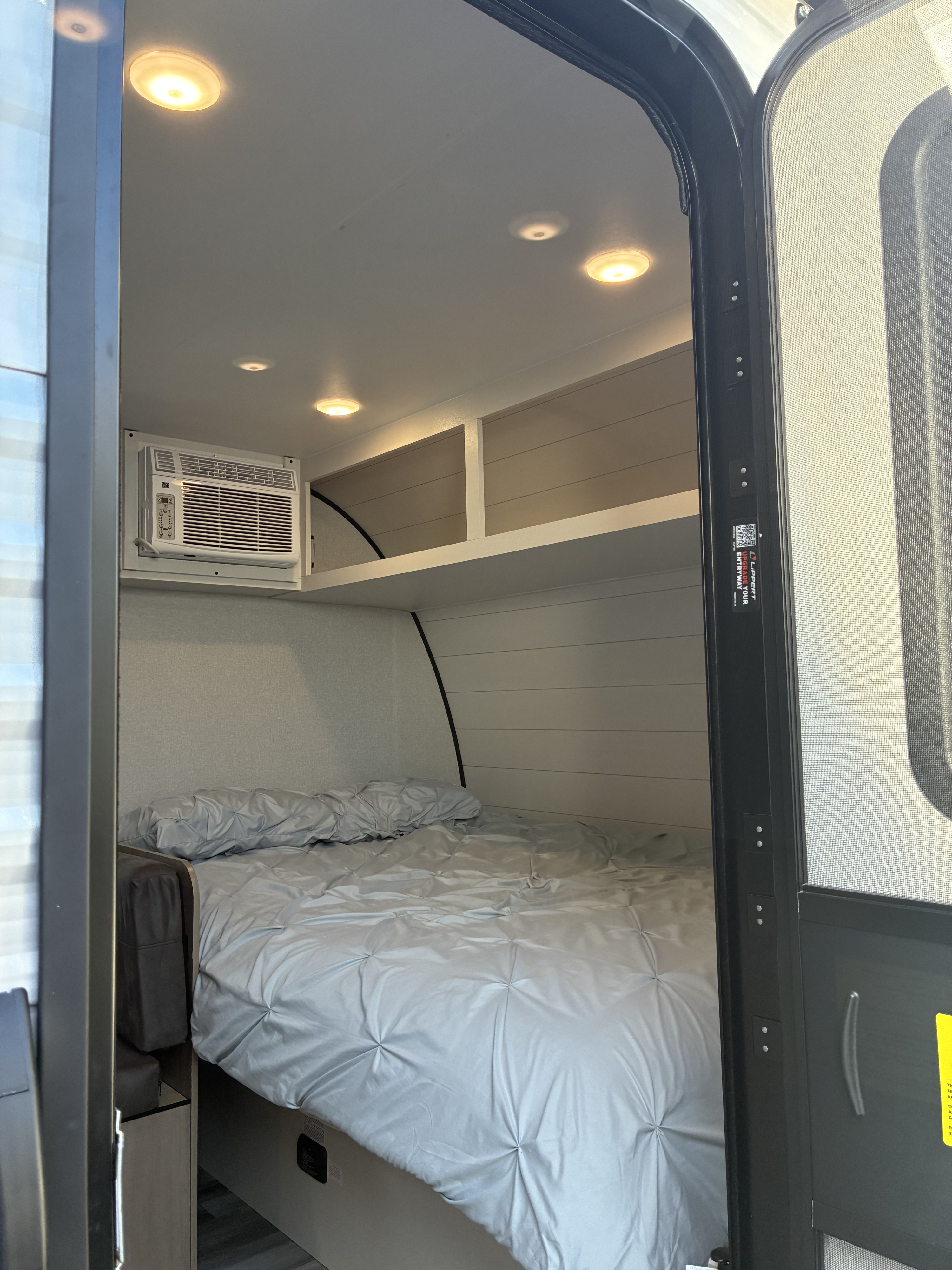 Light-colored RV rental interior featuring a bed with gray ruched bedding, AC unit, and overhead storage, visible from the open door.. Jayco 1UJBC0AJ6F7750334 2026