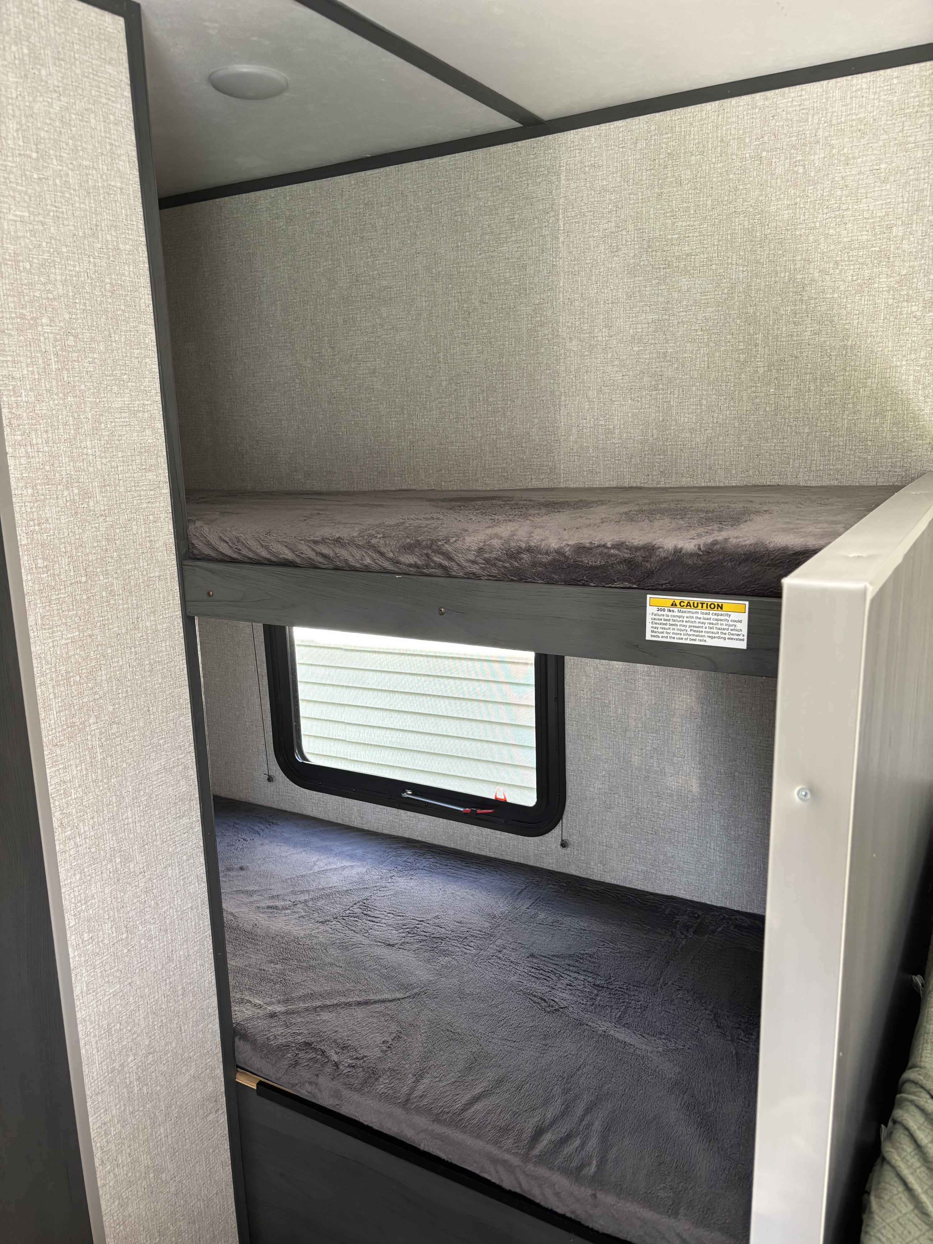 RV rental interior: two bunk beds with grey mattresses, a window, and a clear 300 lbs. max load caution sign.. Dutchmen Aspen Trail 17BH 2022