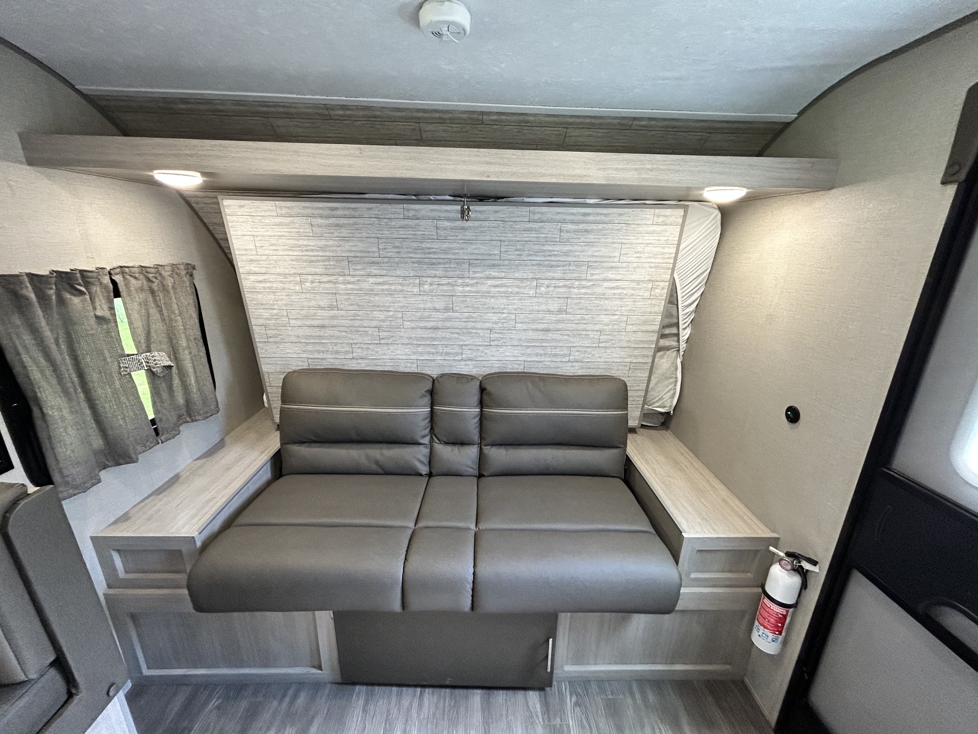 RV rental interior: A modern gray fold-out sofa/bed, overhead storage, window with curtains, and fire extinguisher for safety.. Forest River Aurora Aurora Light 2023