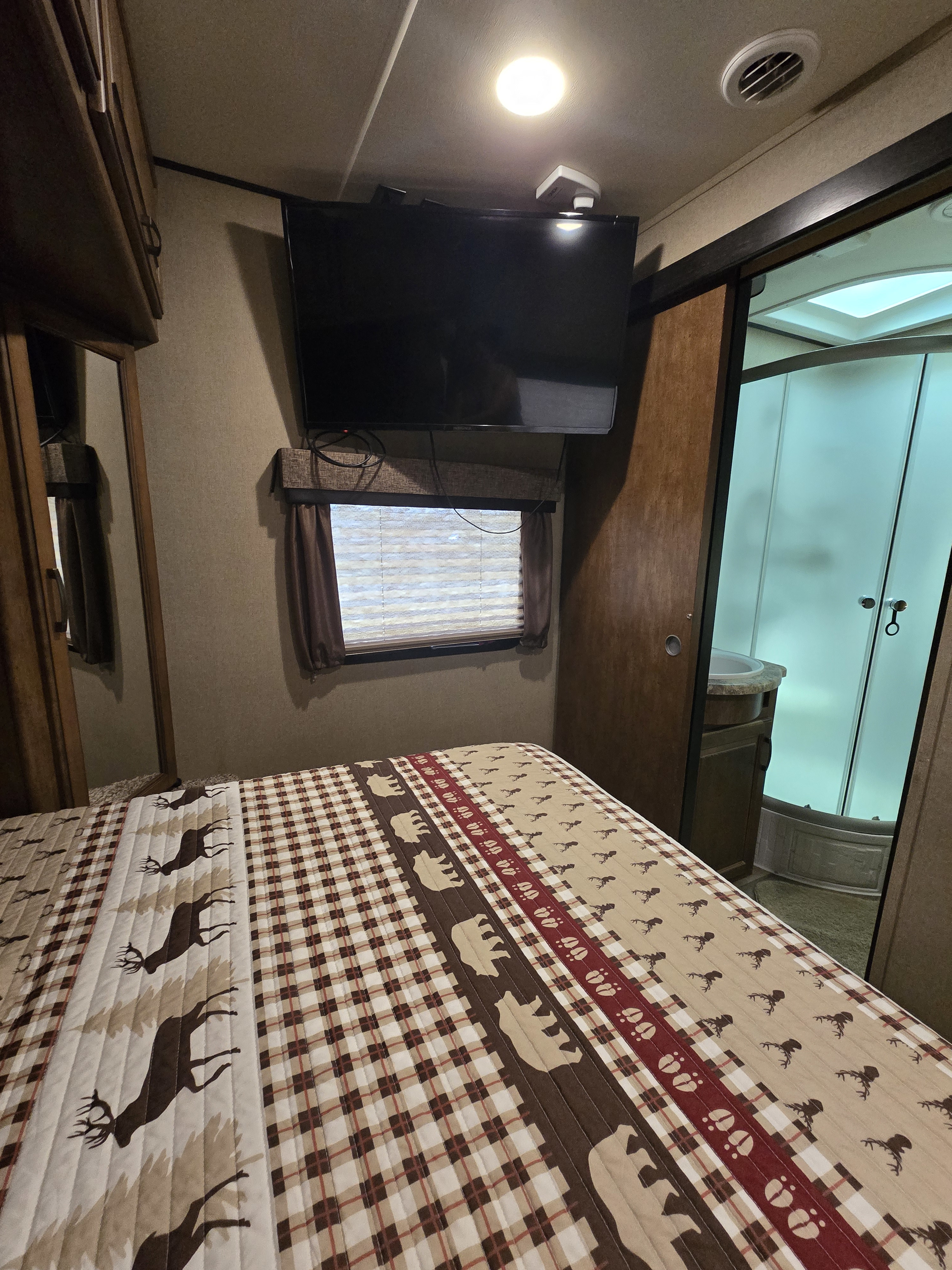 Cozy RV rental bedroom features a plaid and deer-patterned bed, wall-mounted TV, and an adjacent compact bathroom with shower.. Grand Design Reflection 2017