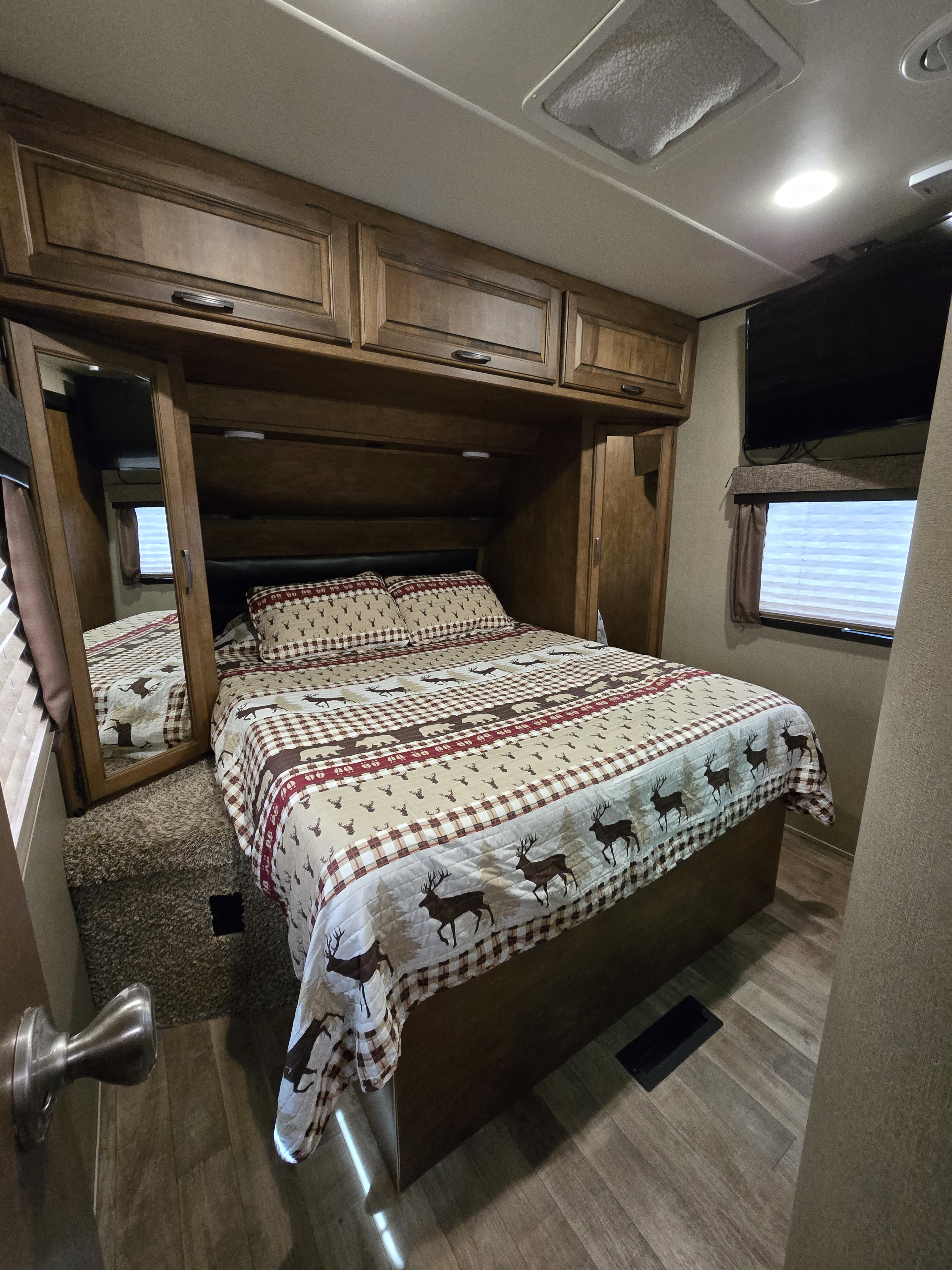 Cozy RV rental bedroom featuring a bed with a deer-patterned comforter, overhead wood cabinets, mirror, TV, and window.. Grand Design Reflection 2017
