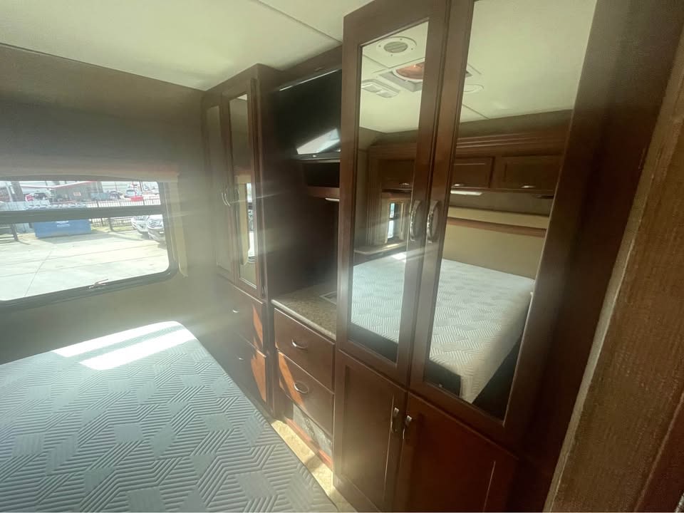 RV rental bedroom with a patterned bed, sunlit window view, and built-in storage, including a mirrored wardrobe and TV.. Thor Motor Coach A.C.E 2015