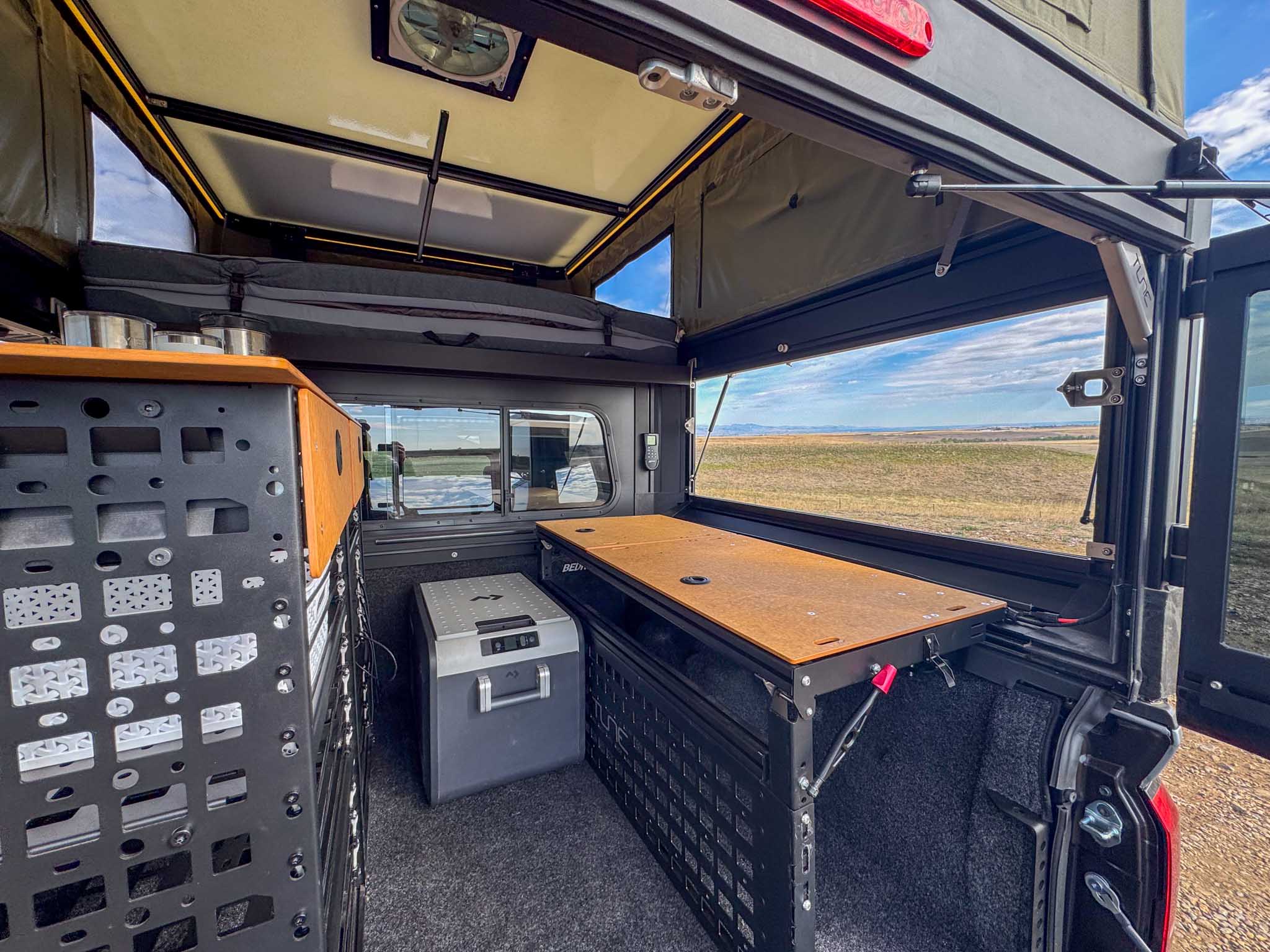 Inside a truck camper, perfect for rv rental. Shows a pop-top tent, fridge, table, storage modules, and an open view of a vast landscape.. Toyota Tacoma 2026
