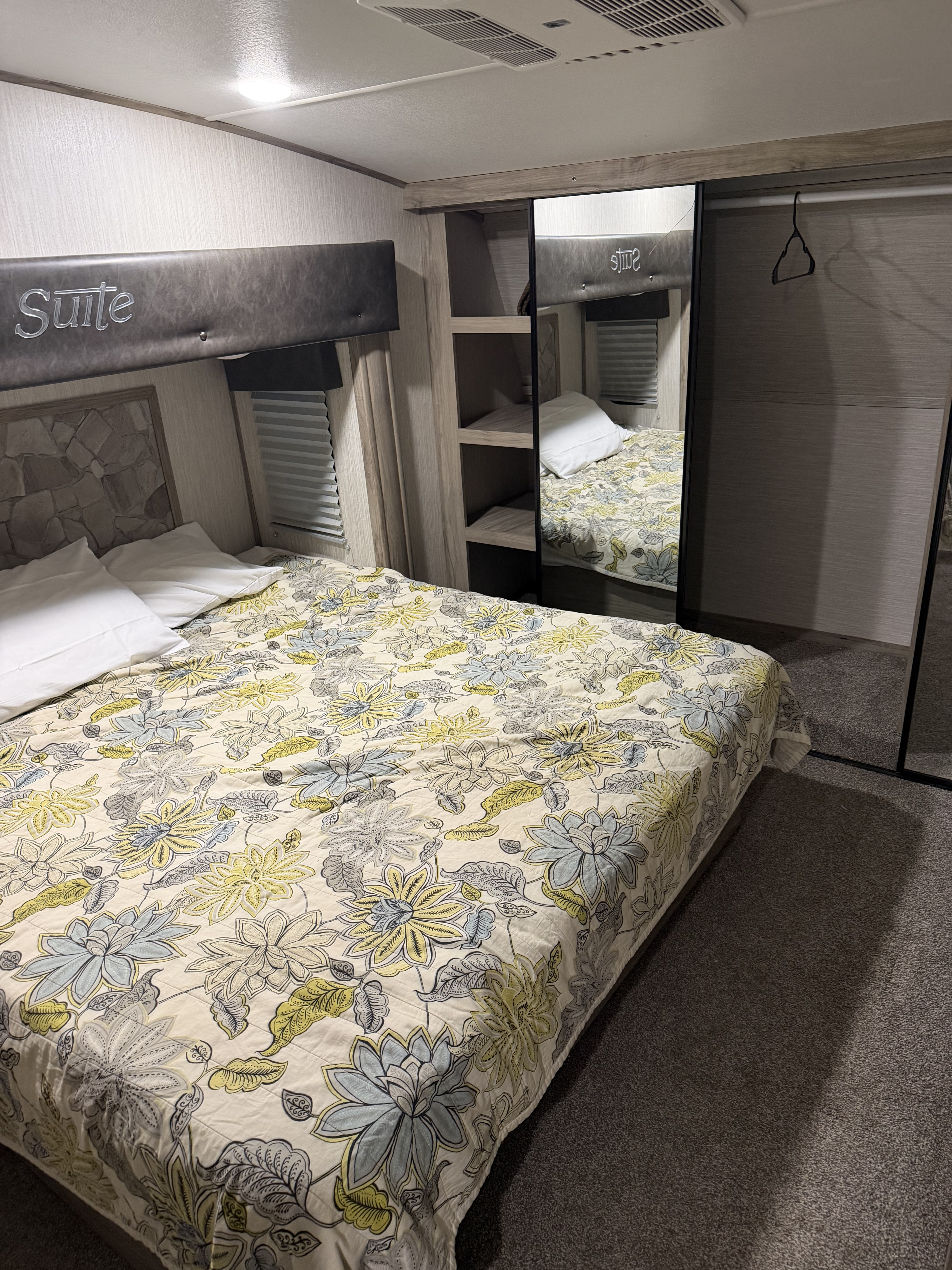 Cozy rv rental bedroom featuring a floral bedspread, "Suite" headboard, and mirrored closet with a hanger.. Forest River Cherokee Arctic Wolf 2020