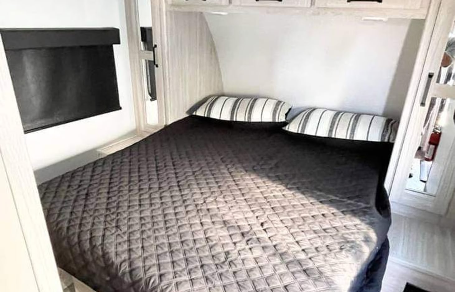 Cozy RV rental bedroom featuring a large bed, striped pillows, dark quilted blanket, and window blind.. Heartland RVs Sundance Ultra Lite 2022
