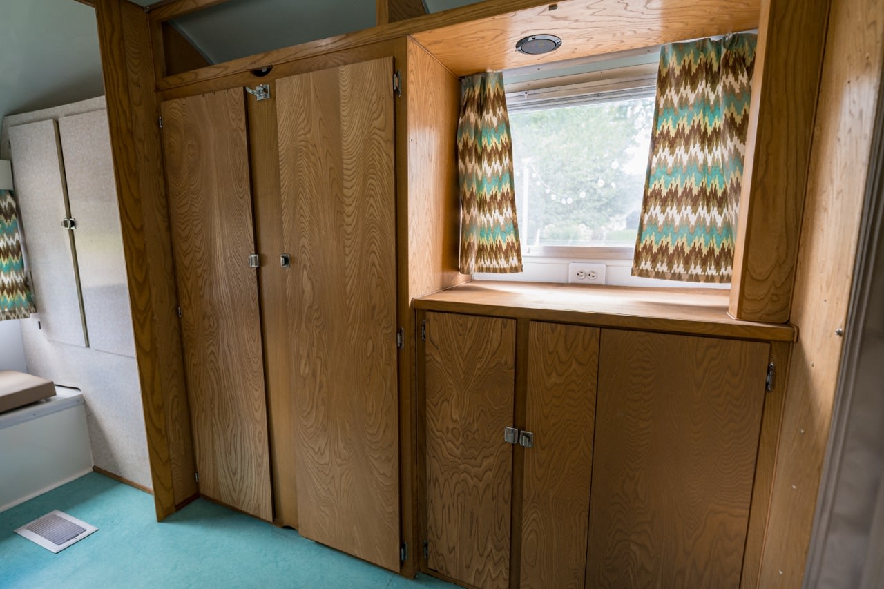 This RV rental interior offers light wood cabinetry, a window with retro patterned curtains, and a compact living space.. Airstream Tradewind 1966