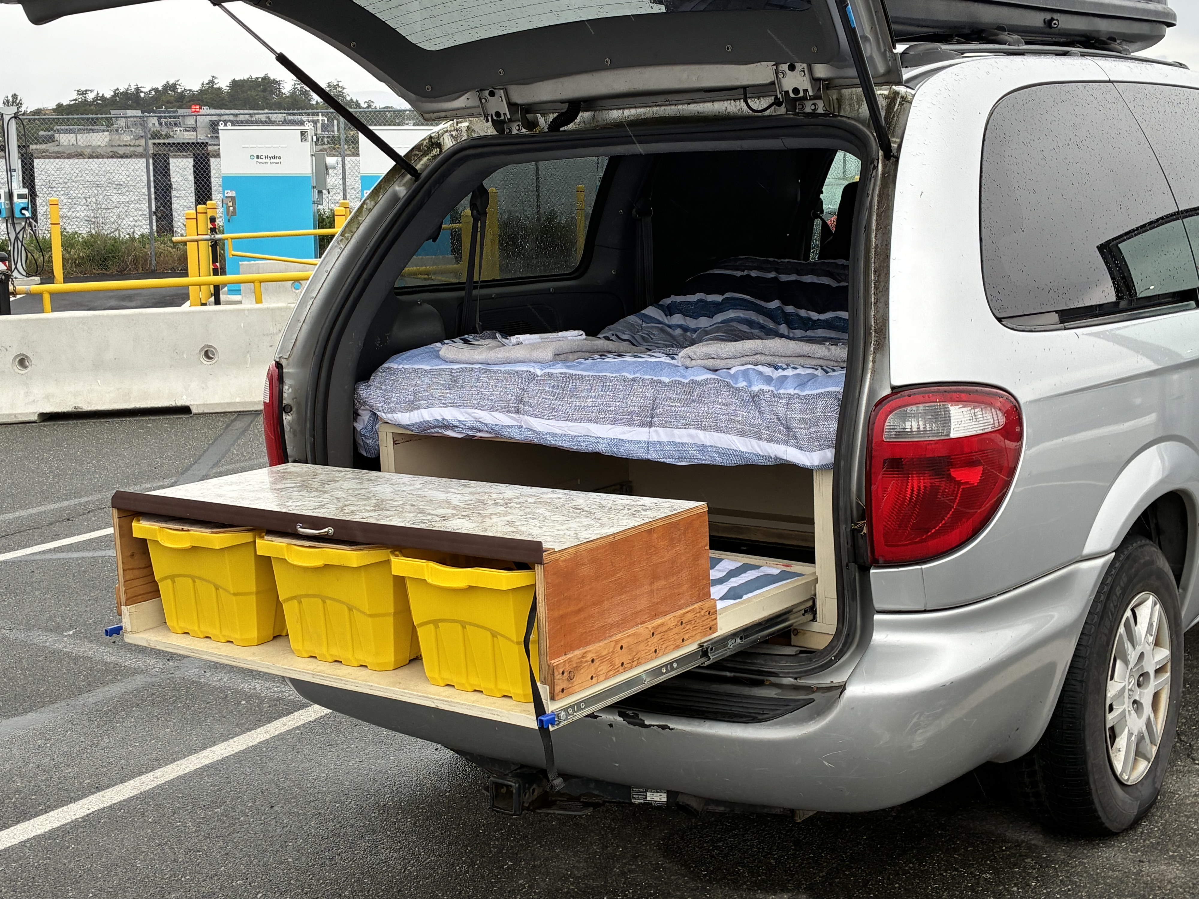 Silver minivan RV rental setup: rear hatch open reveals a cozy bed inside and pull-out storage with yellow bins, ready for adventure.. Dodge Grand Caravan 2005
