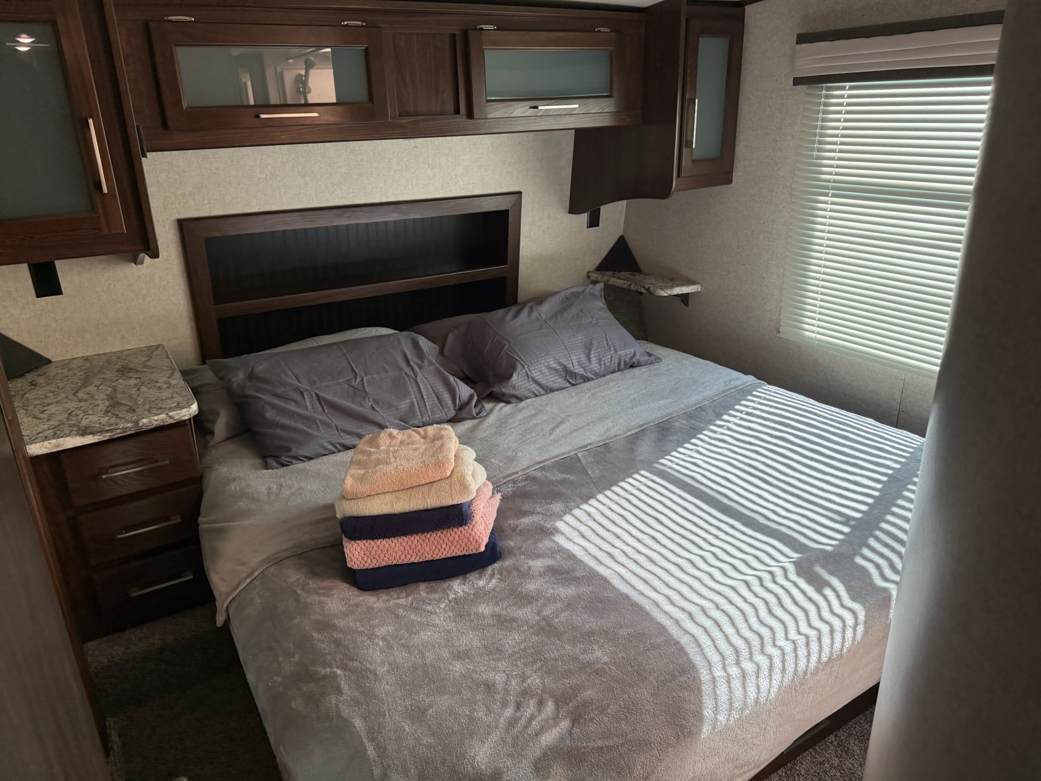 Cozy RV rental bedroom with a comfortable bed, stacked towels, wooden cabinets, and sunlit window blinds.. Heartland RVs Torque Toy Hauler 2017