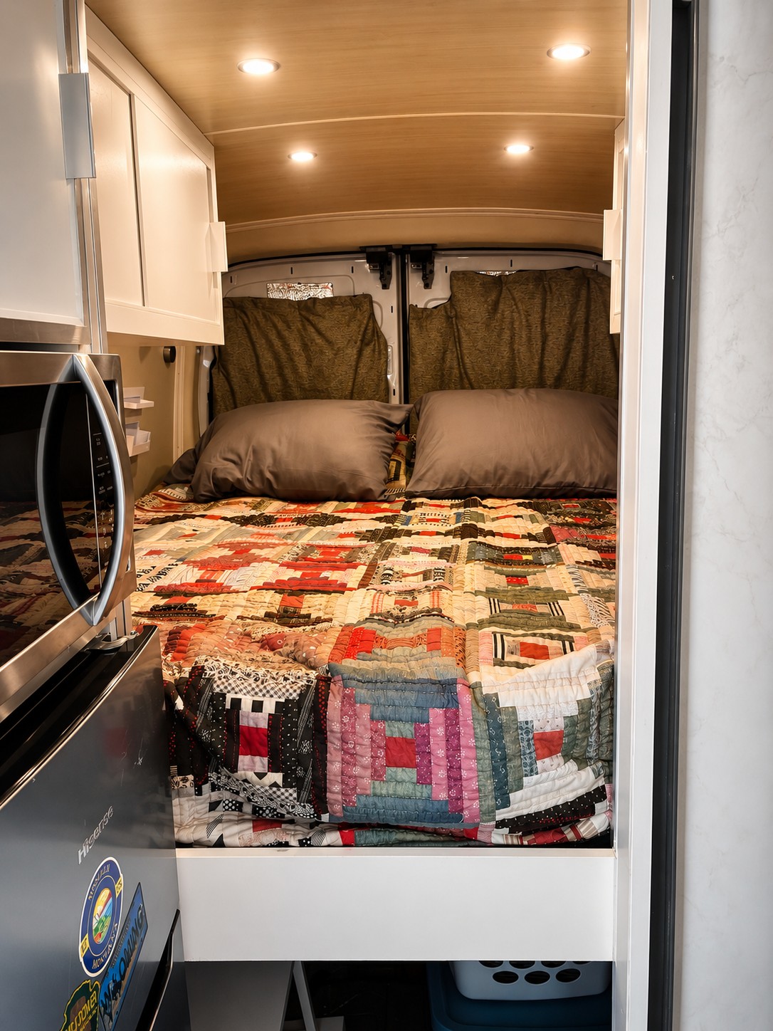 Inside a cozy RV rental: a colorful patchwork bed, wood ceiling, and compact kitchen with white cabinets and a microwave.. Ford F350 2019