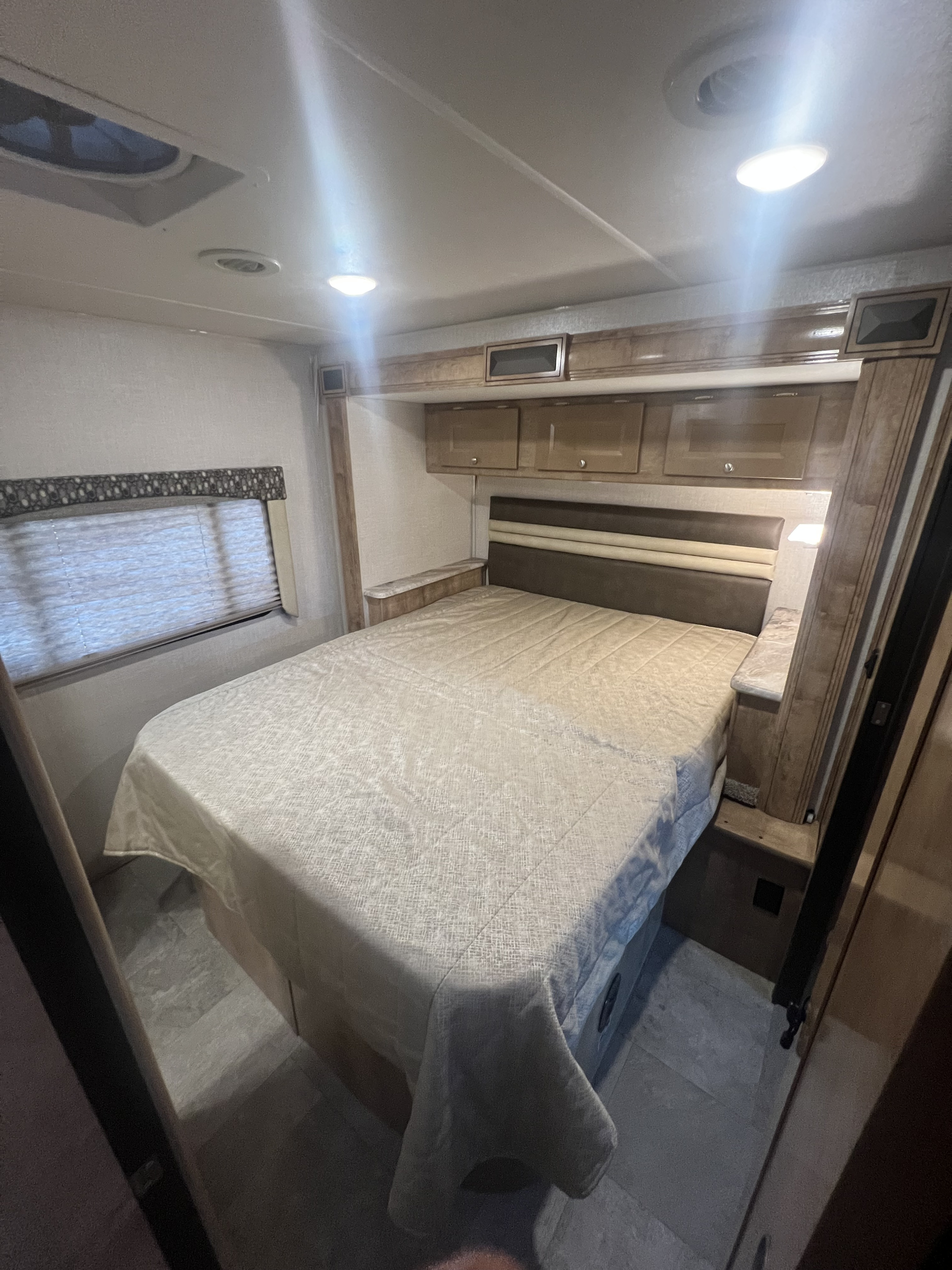 A view inside an RV rental's bedroom, showcasing a neatly made bed, overhead storage, a window, and wood-toned cabinetry.. Forest River Coachmen Leprechaun 2020