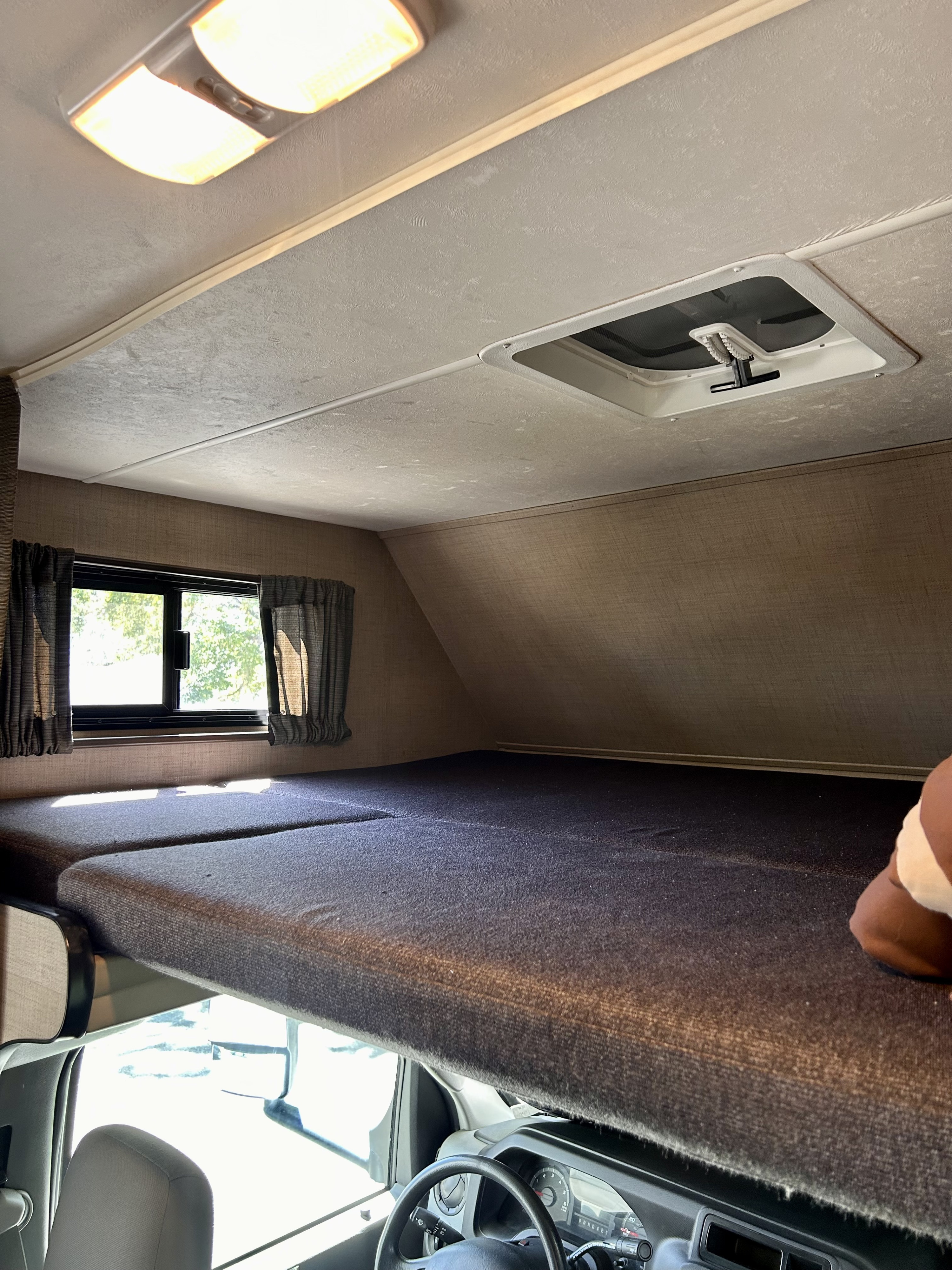 Inside an RV rental: a bright, cozy overhead bunk sleeping area with window, ceiling light, and vent, above the driver's cab.. Thor Majestic 2018