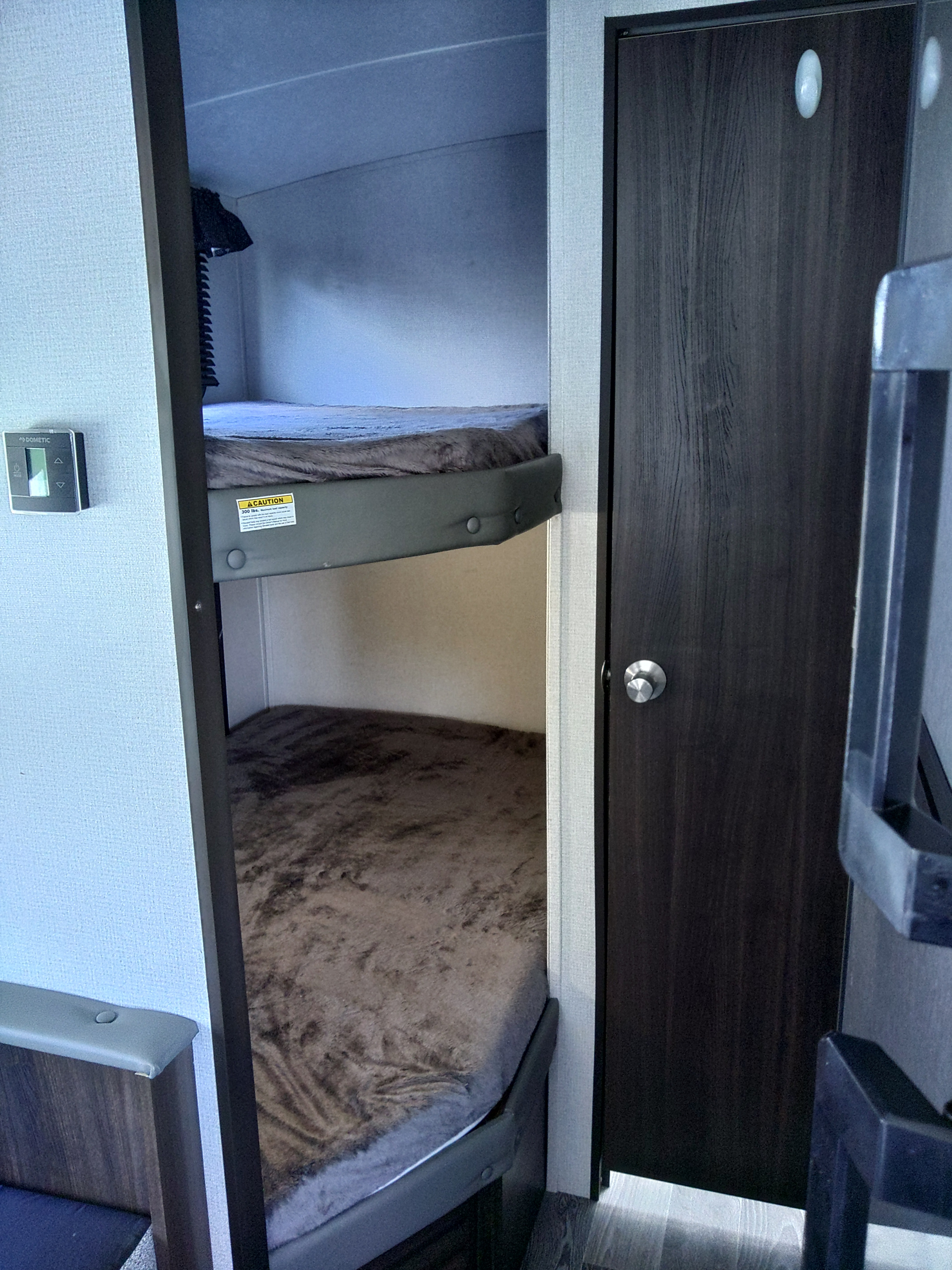 Inside an RV rental, two bunk beds with cozy blankets are visible. A Dometic thermostat is on the left wall, and a dark door is on the right.. Keystone RV Springdale 2020