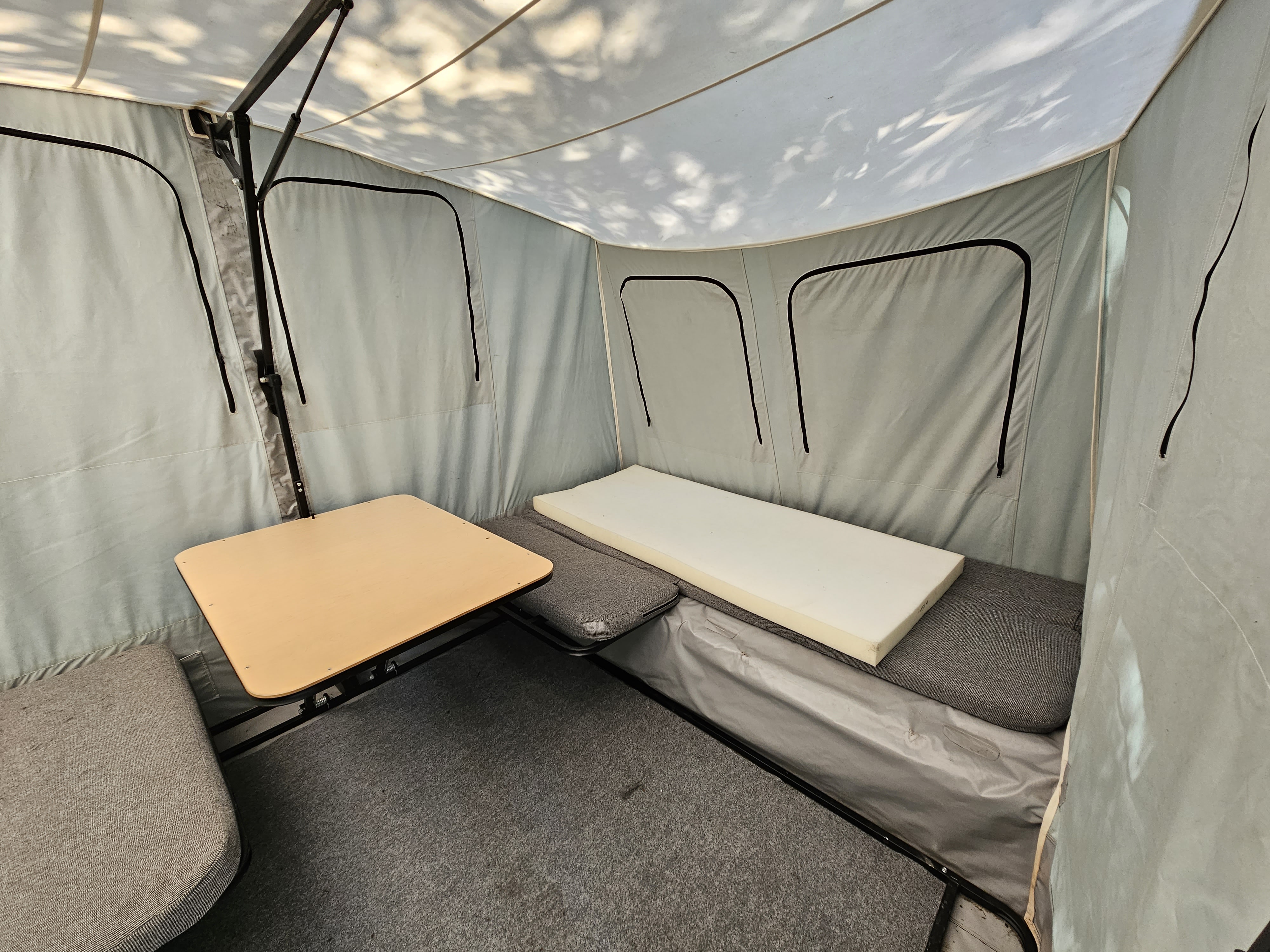 Inside an RV rental pop-up, showing a small table, grey cushioned seating, a foam bed, and carpeted floor.. Jumping Jack 6x8 REGULAR 2012