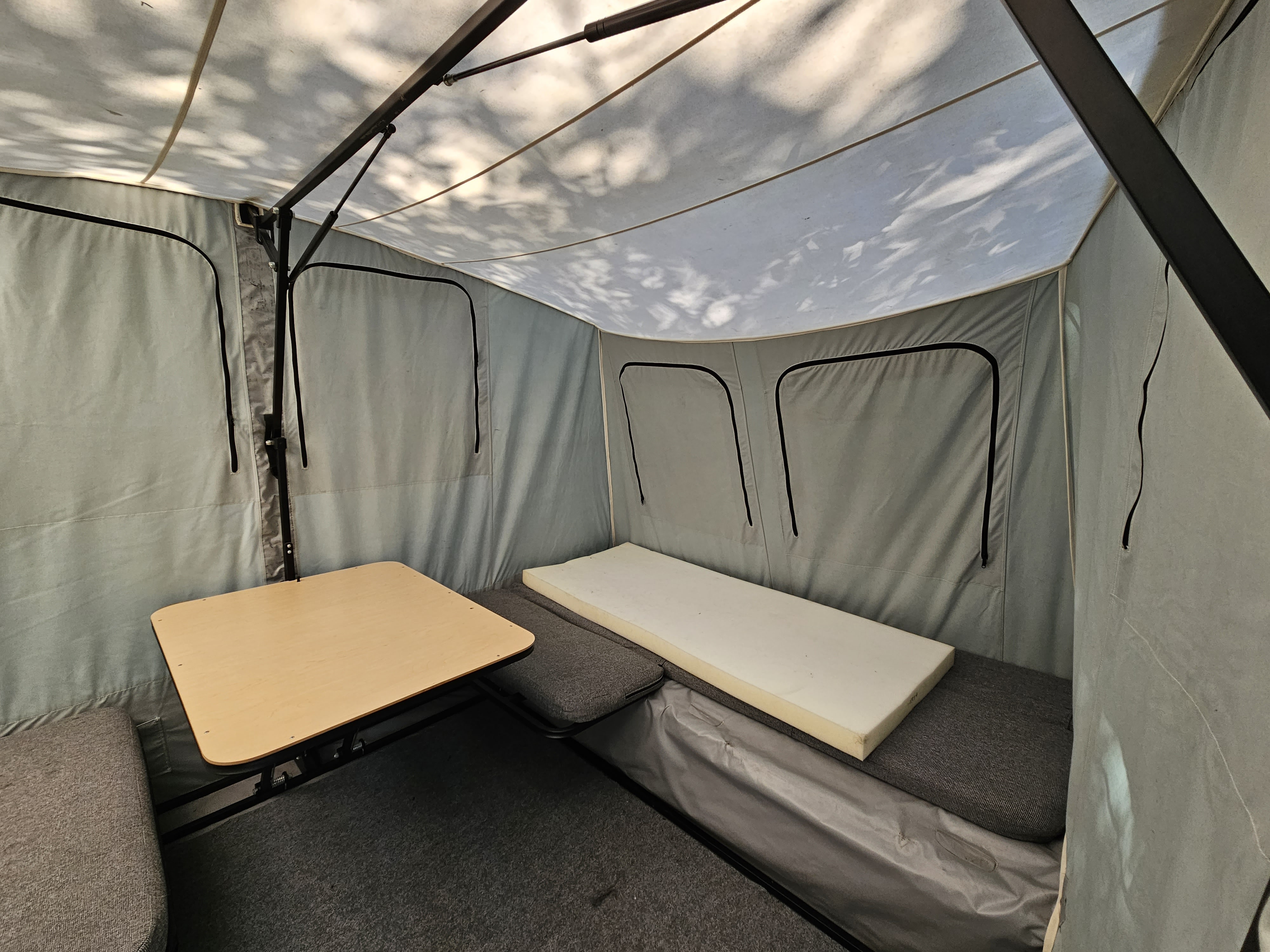 Inside an RV rental camper: light grey canvas interior with a wooden table, grey cushions, and a foam mattress.. Jumping Jack 6x8 REGULAR 2012