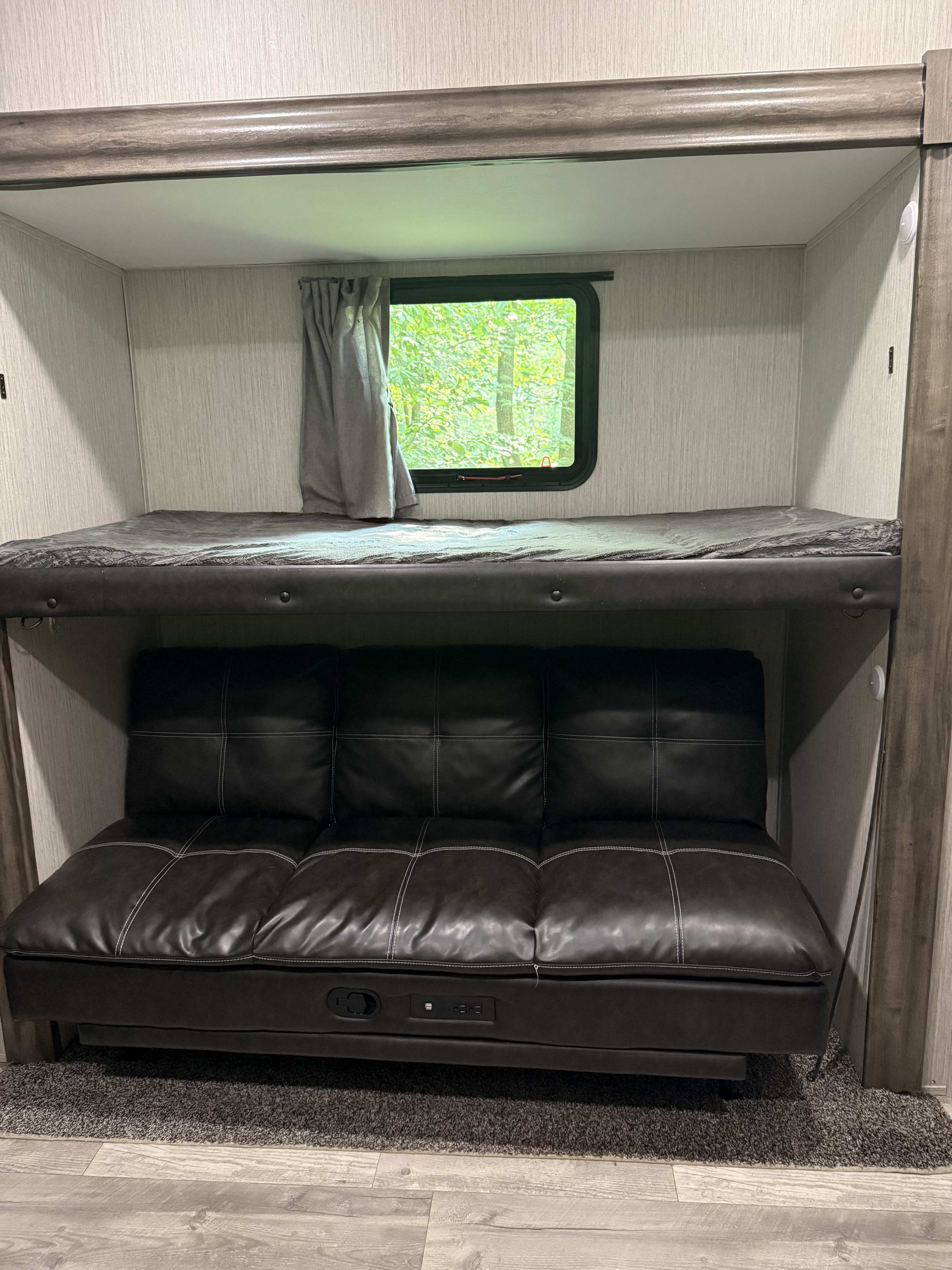 This RV rental features a bunk bed above a futon with USB charging ports. A window overlooks green trees.. Forest River Sandpiper 2021