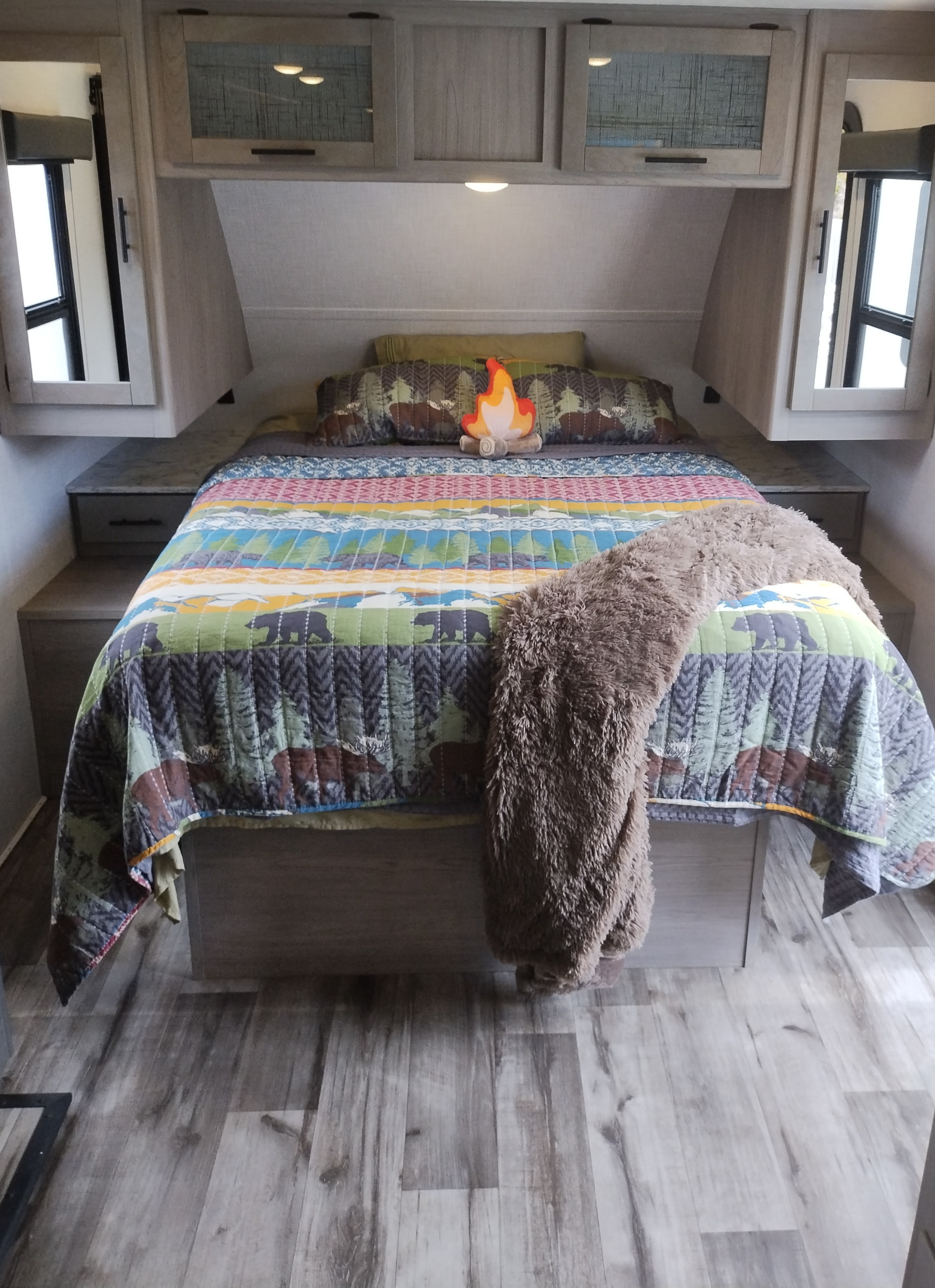 An inviting RV rental bedroom featuring a bear and moose quilt, plush fire pillow, overhead cabinets, and side windows.. Dutchmen Coleman Light 2023