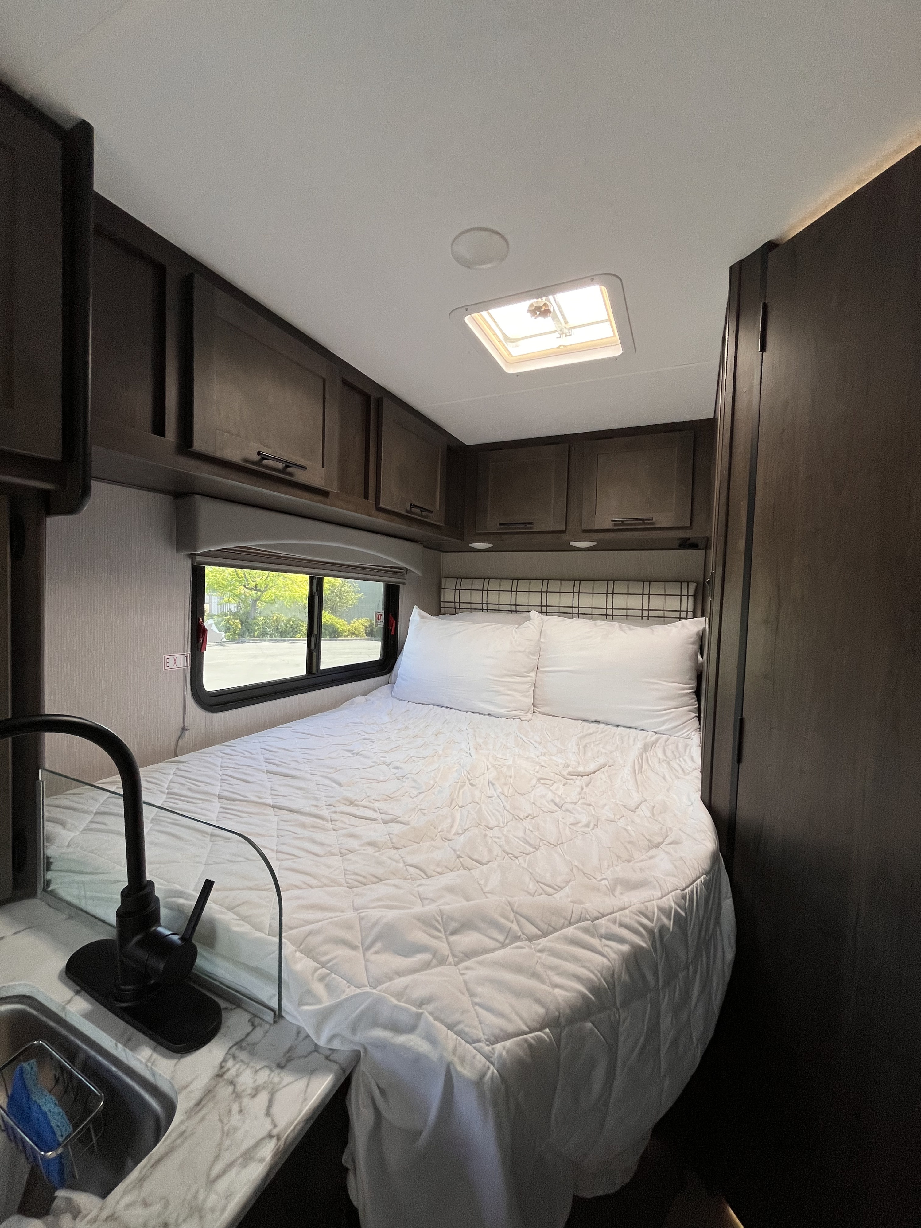 Cozy RV rental bedroom featuring a white bed, plaid headboard, sink, and overhead storage. An ideal compact living space.. Coachmen Cross Trail 2023