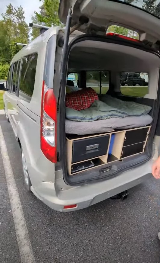 Inside a compact RV rental camper van: bed, sleeping bag, and custom storage with cooler visible from the open tailgate.. Ford Transit 2016