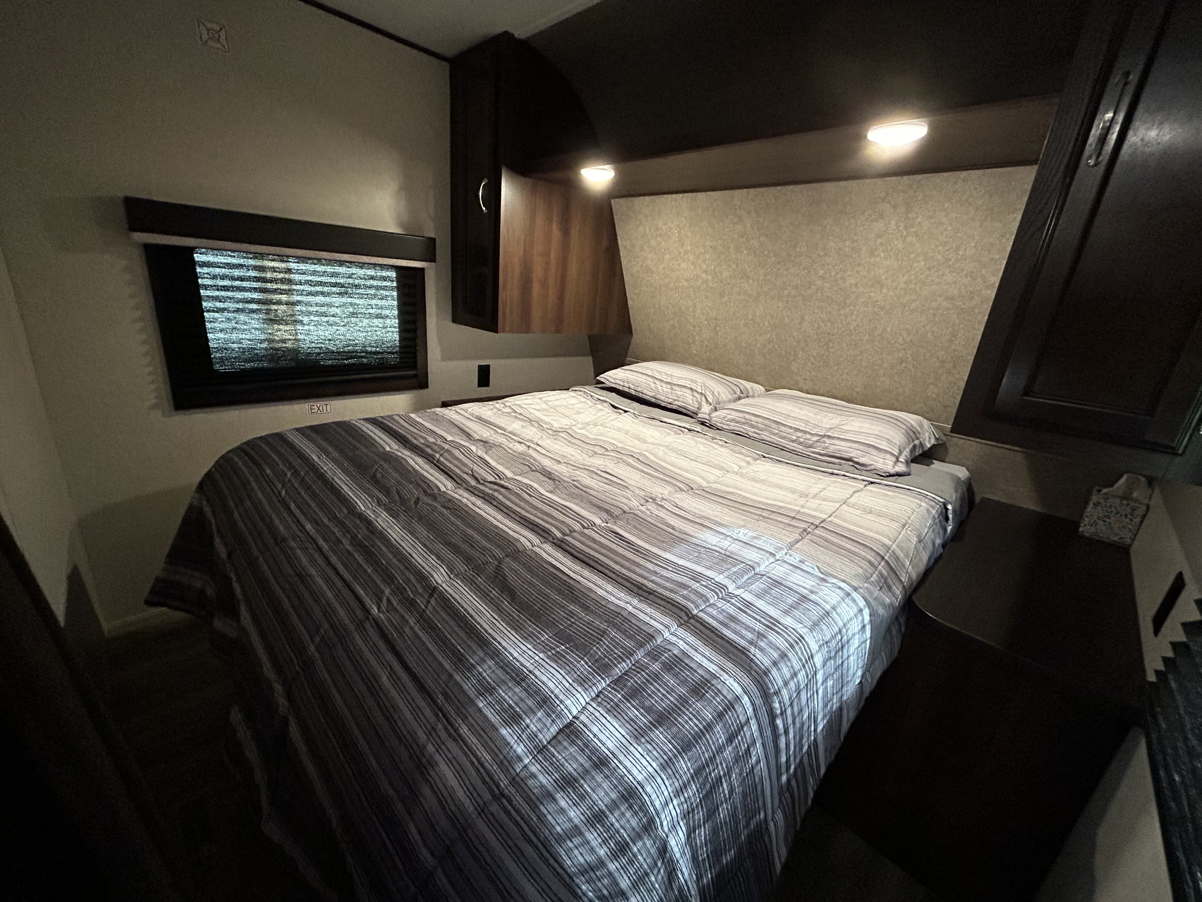 A compact RV rental bedroom features a striped bed, bright reading lights, overhead storage, and a window with an 'EXIT' sign.. Jayco Jay Flight SLX 2018